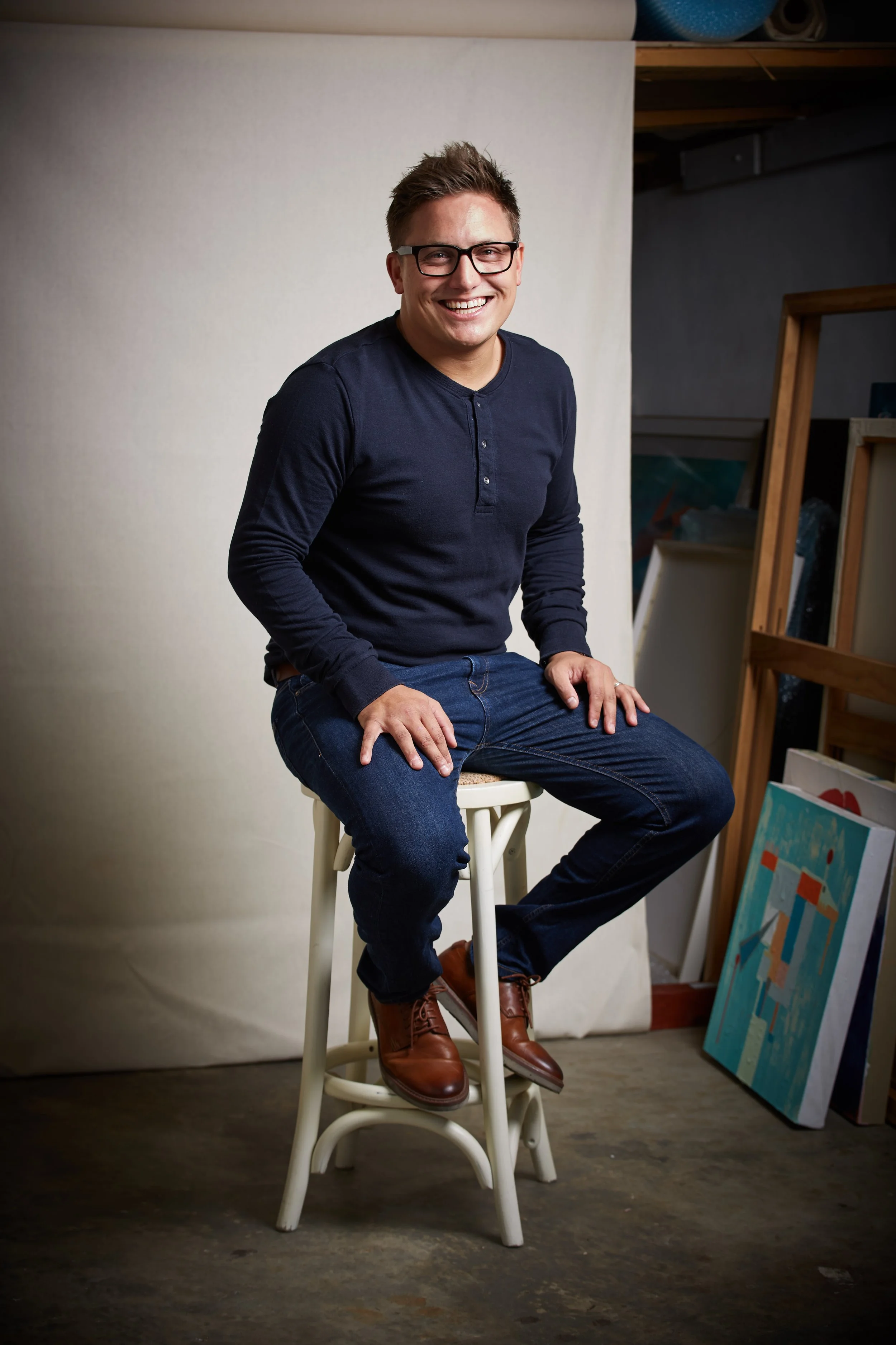 A man with glasses and a navy blue long-sleeve shirt sitting on a white stool in front of a plain light-colored backdrop, with art supplies and paintings visible in the background.