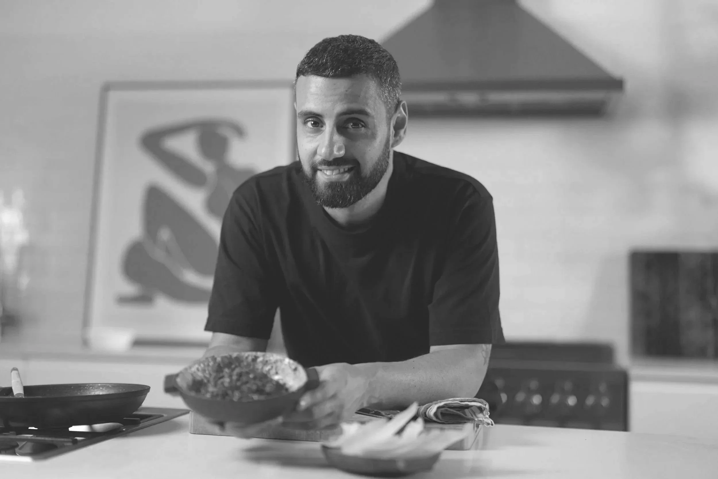 A man with a beard and short hair smiling while preparing food in a kitchen. There are bowls, utensils, and food on the counter.