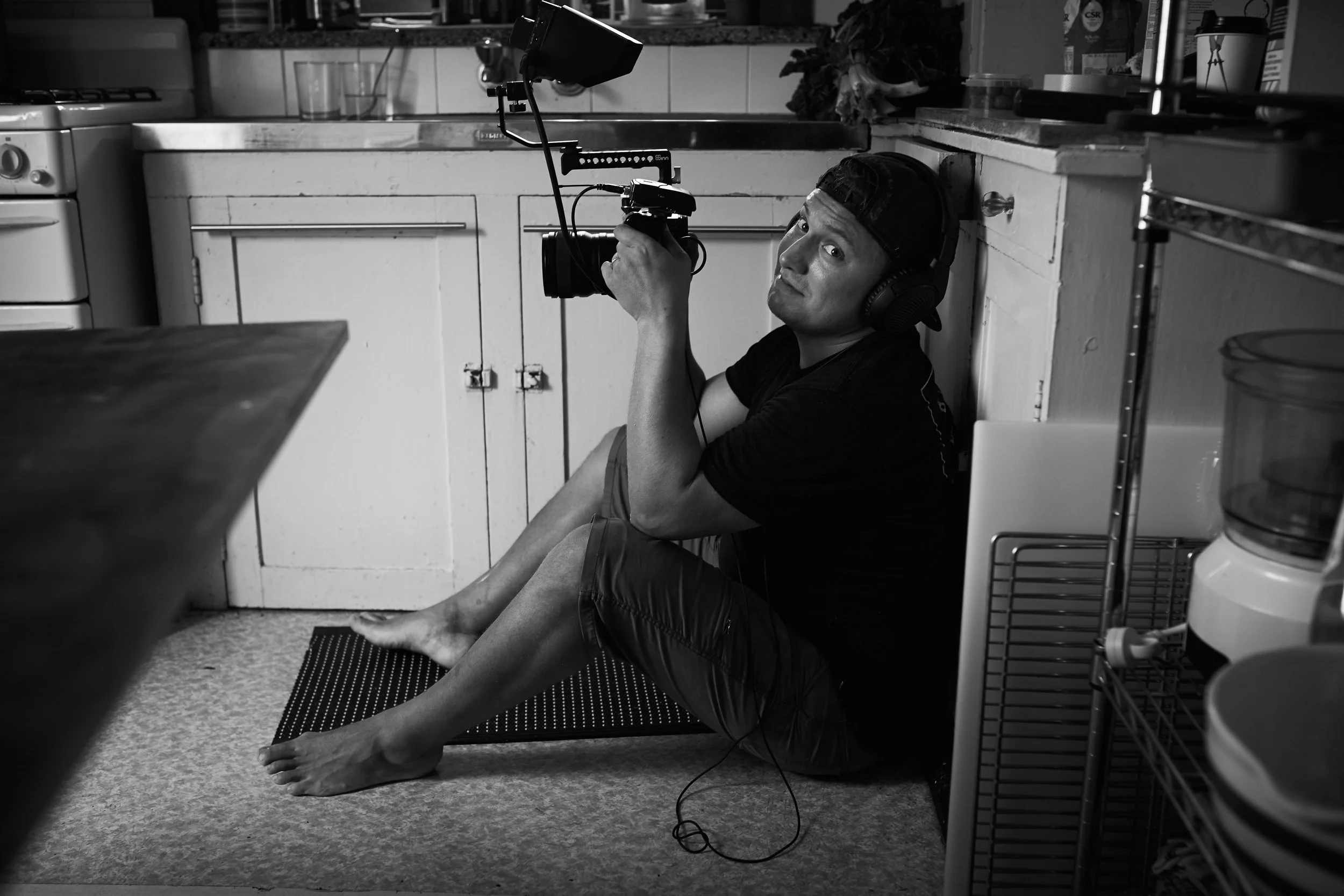 A man lying on his back on the floor of a kitchen, holding a camera and looking up at the lens. He is wearing headphones and a backward cap, with his feet bare and resting on a rug.