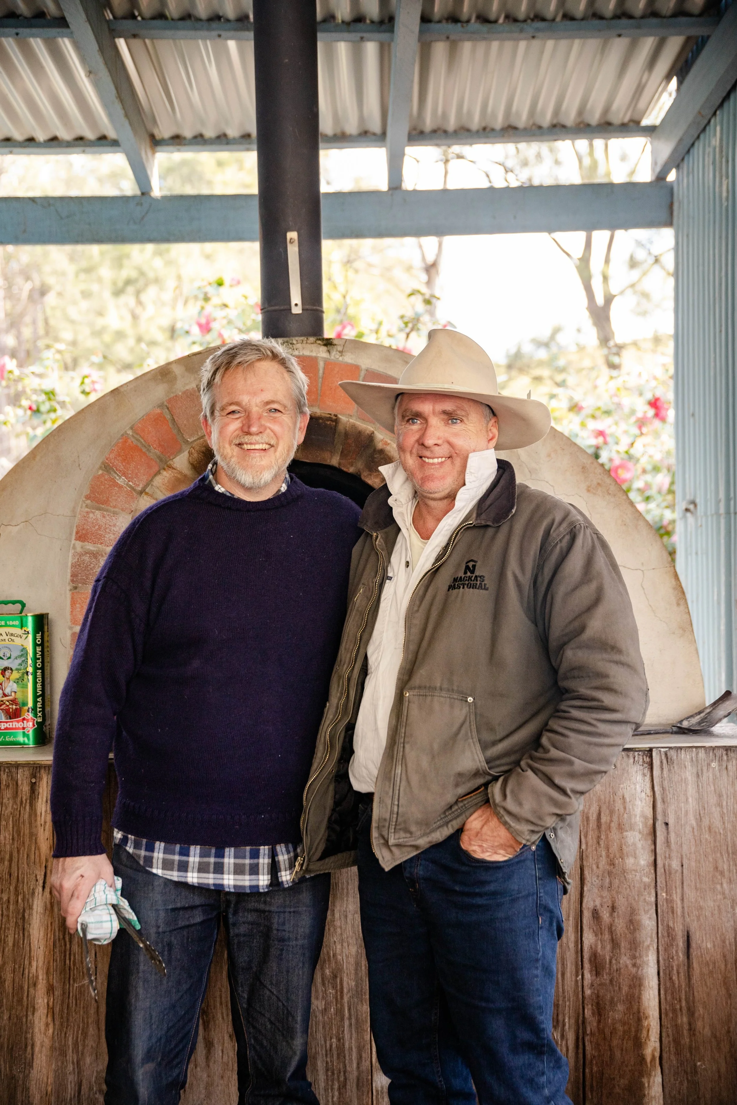 Two smiling men stand together in front of a brick oven, one wearing a dark blue sweater and plaid shirt, holding a cloth, the other wearing a wide-brimmed hat and a gray jacket, under a corrugated metal roof with trees and flowers in the background.