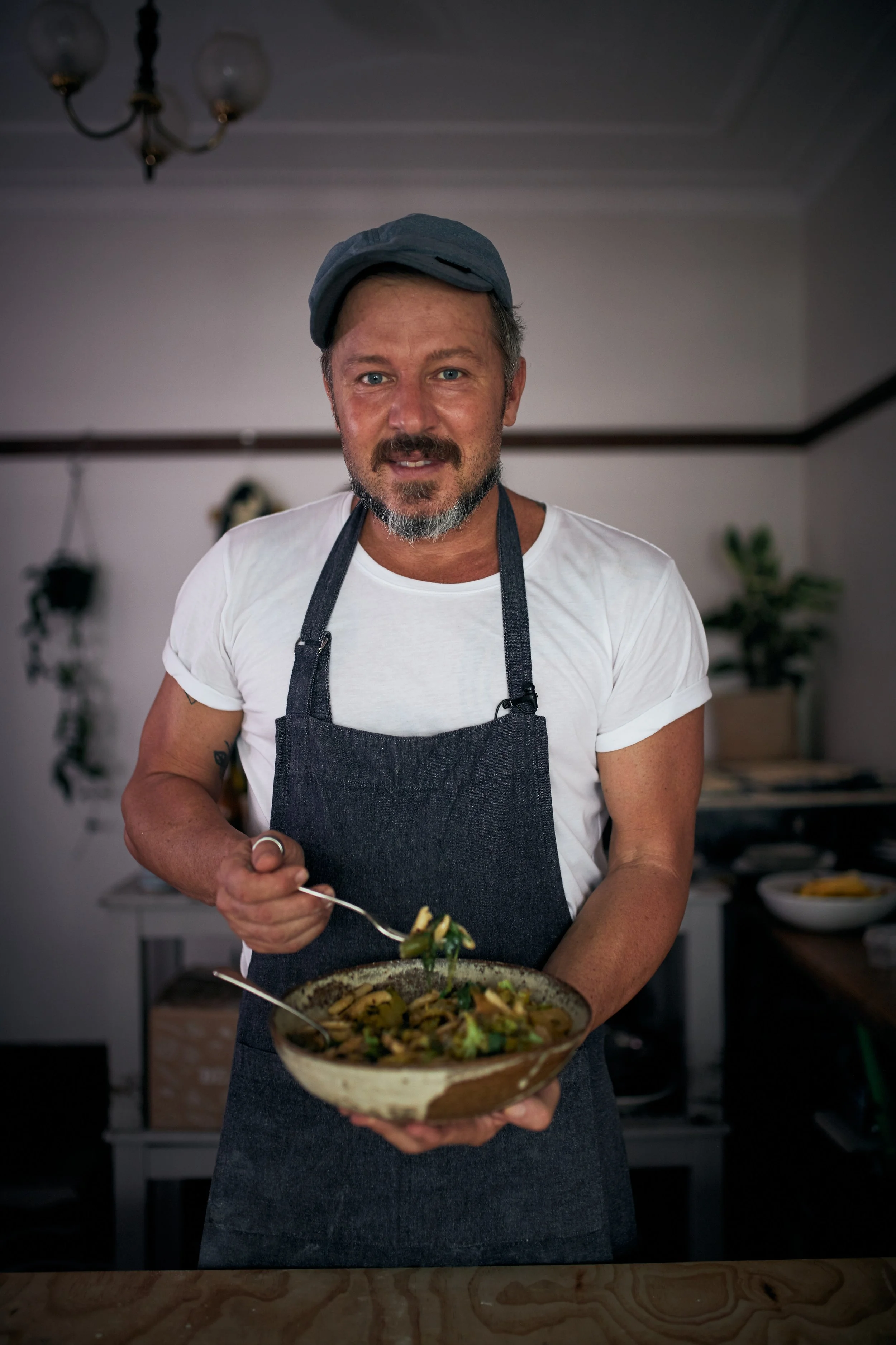 A man with a beard and blue eyes wearing a gray cap and white t-shirt, holding a bowl of food with a spoon, standing in a kitchen.