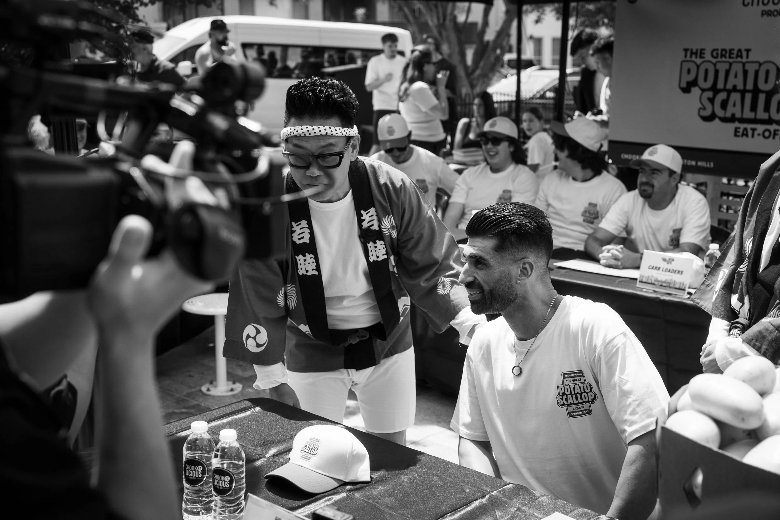 People gathered around a table at an outdoor event, with some wearing hats and t-shirts advertising 'The Great Potato Scalopp'. A man with glasses and a polka-dot headband is in focus, and there are water bottles and caps on the table.