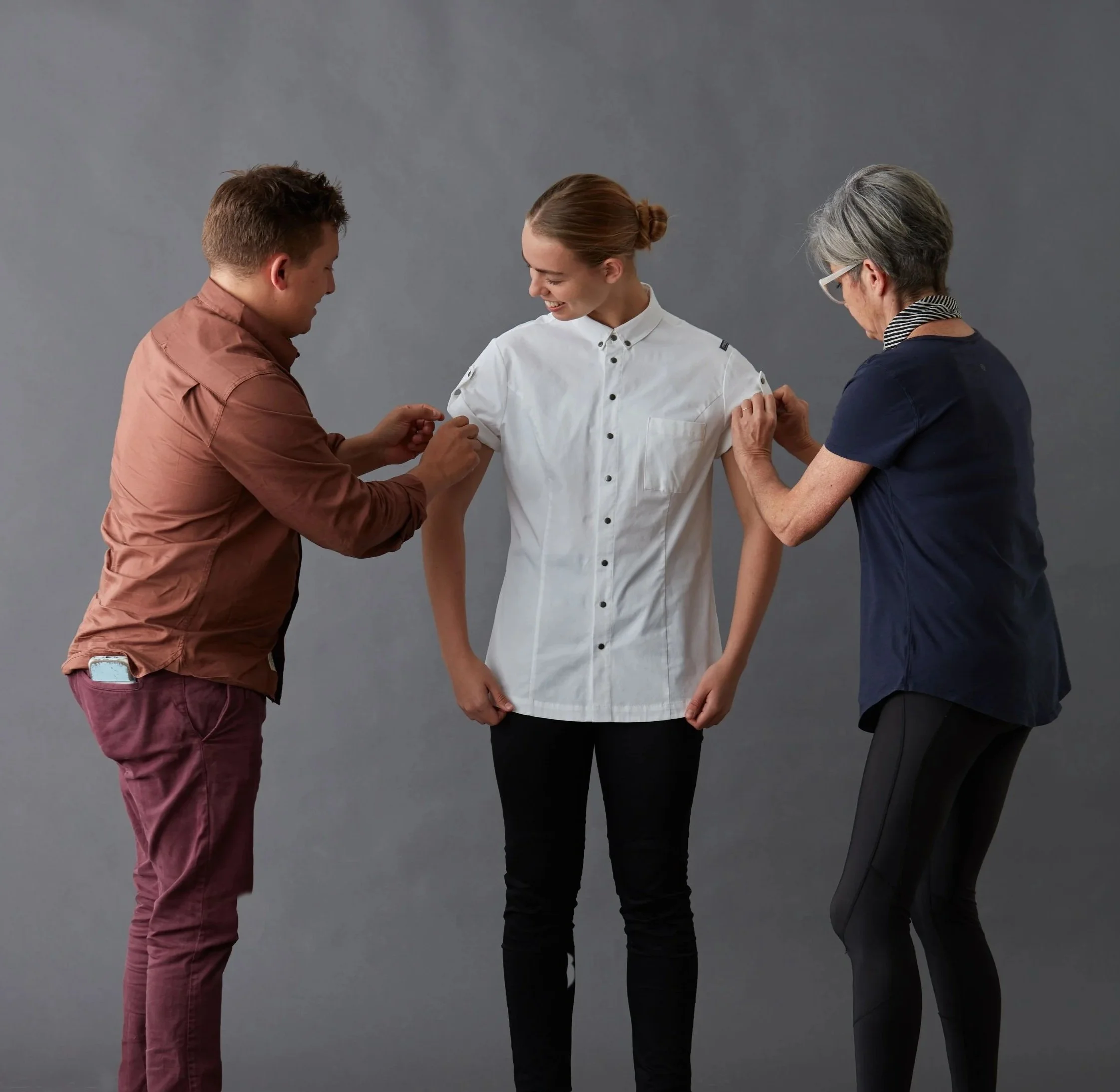 Three people, one young man, one young woman, and one older woman, are engaged in a friendly interaction against a plain gray background. The young man and young woman are smiling and appear to be sharing a happy moment, while the older woman, wearing glasses, is looking at the interaction.
