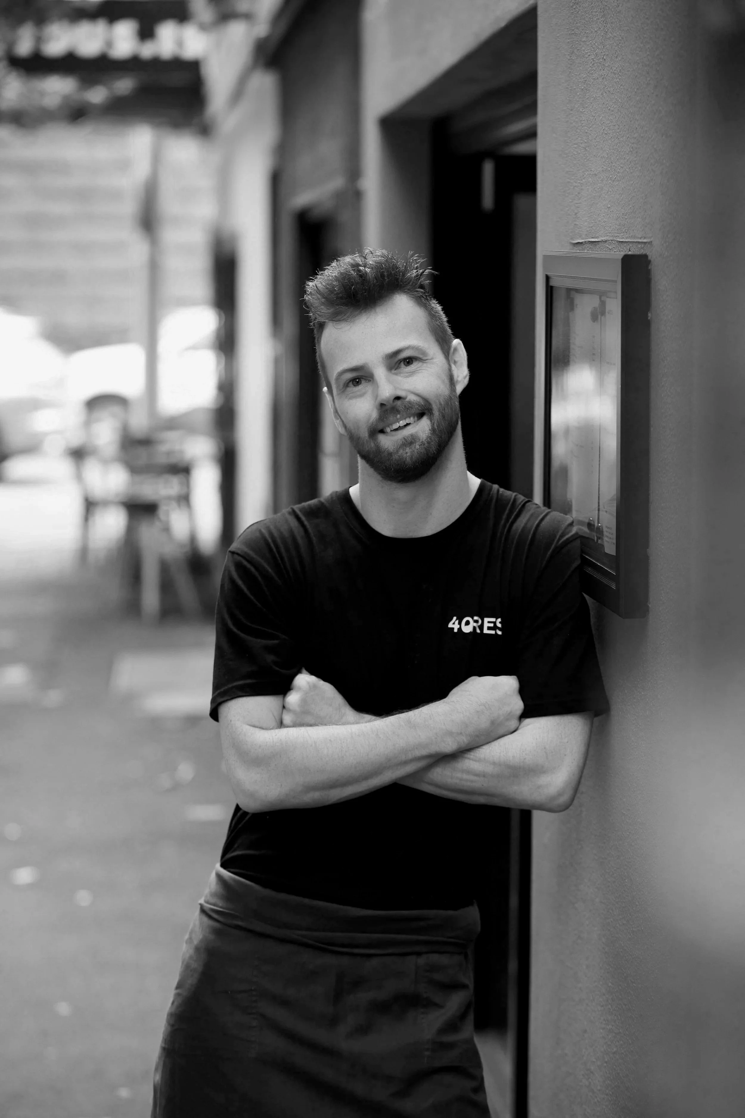 A man with a beard and short hair leaning against a wall outside, smiling with his arms crossed, wearing a black t-shirt with a logo, in an urban setting.