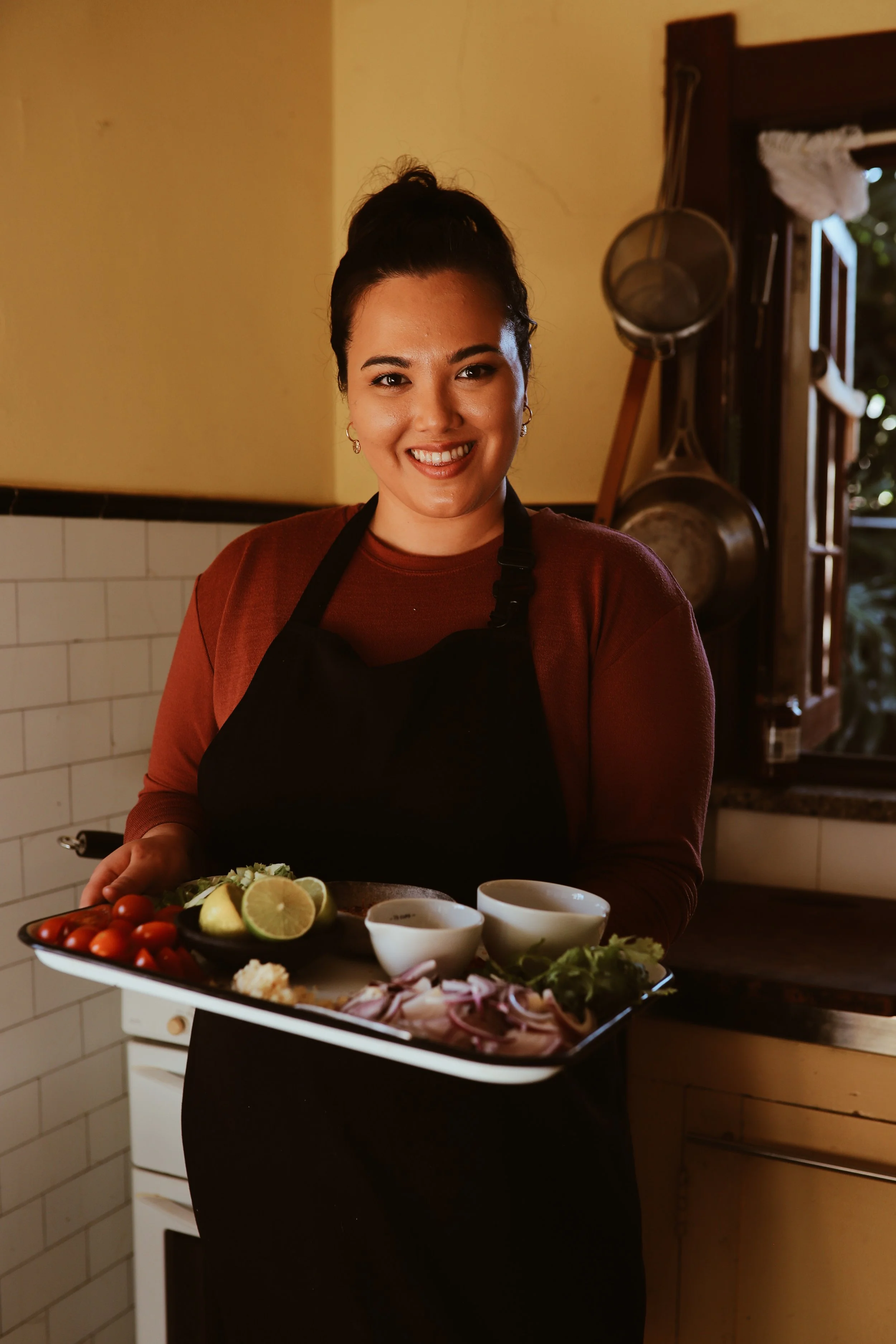 A woman in a kitchen holding a tray of fresh vegetables and lime slices, smiling at the camera.