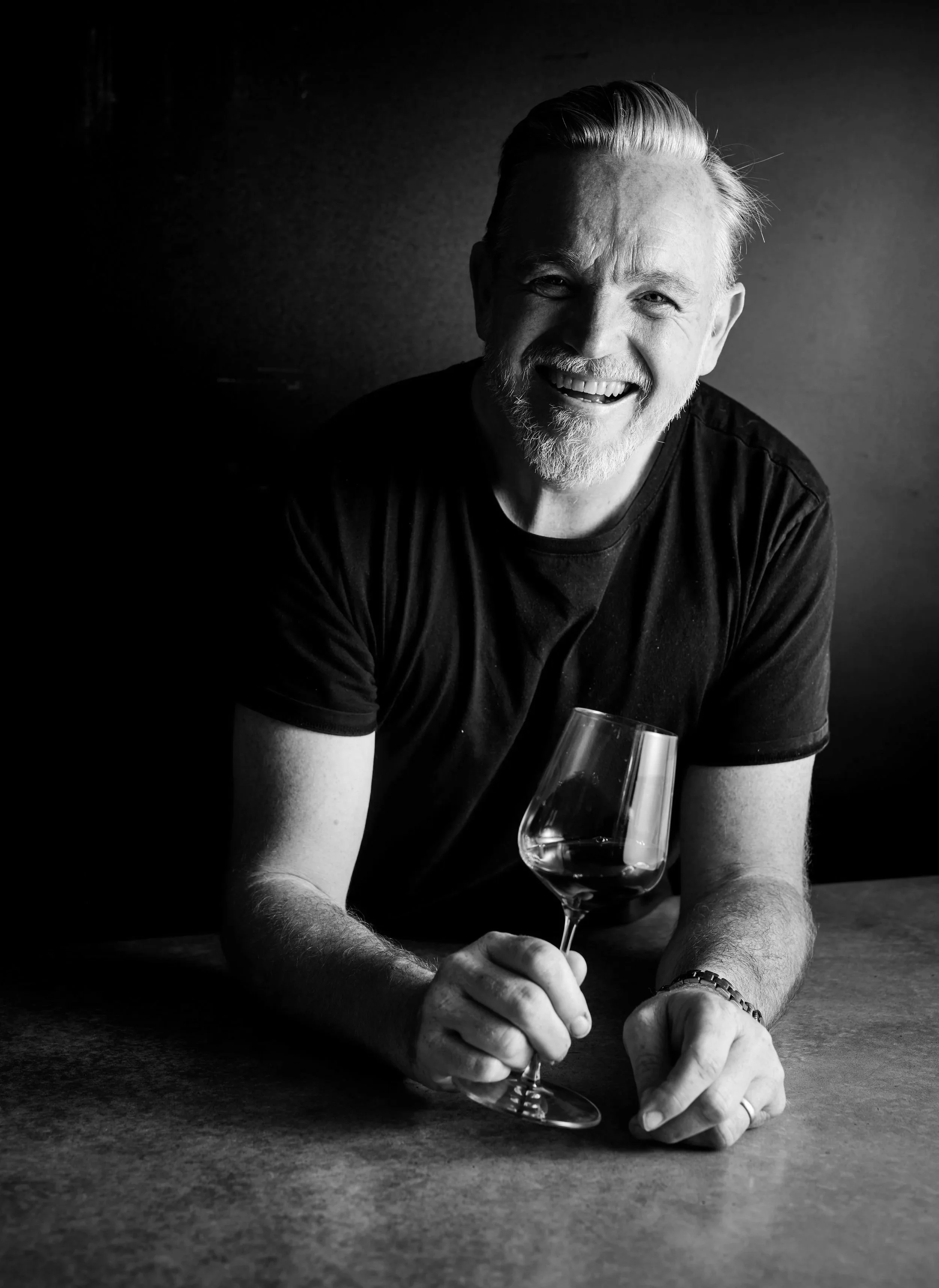 A smiling man with a beard and mustache, wearing a black T-shirt, is sitting at a table holding a wine glass in one hand. The photo is black and white, with dramatic lighting highlighting his face and hands.