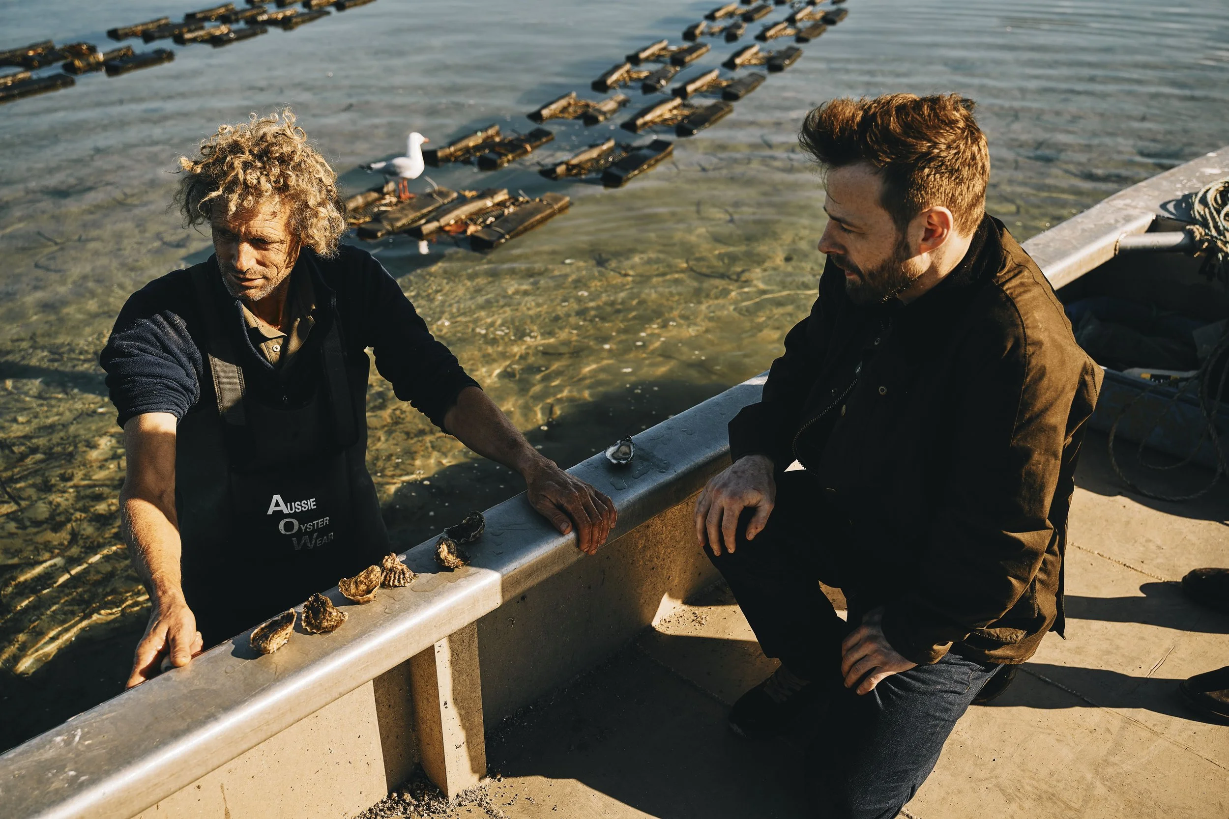 Two men are sitting by the water, examining shells or rocks on a concrete curb near the shoreline. One man has curly gray hair and is wearing a black apron with the words 'Aussie Oyster Wear,' while the other man has short brown hair and is wearing a black jacket. A seagull is standing on the water in the background, and there are wooden objects floating on the water.