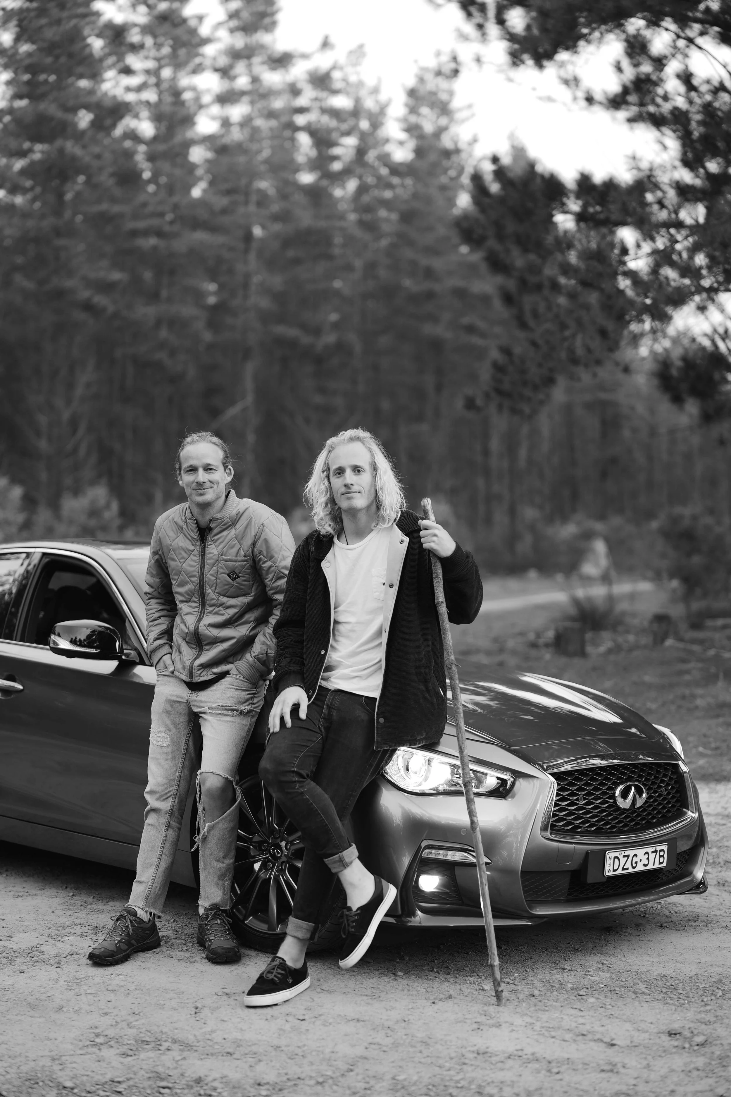 Two men standing and leaning against a sleek, black Infiniti car in an outdoor setting with trees in the background. One man is wearing a jacket and the other is dressed in a hoodie, holding a walking stick.