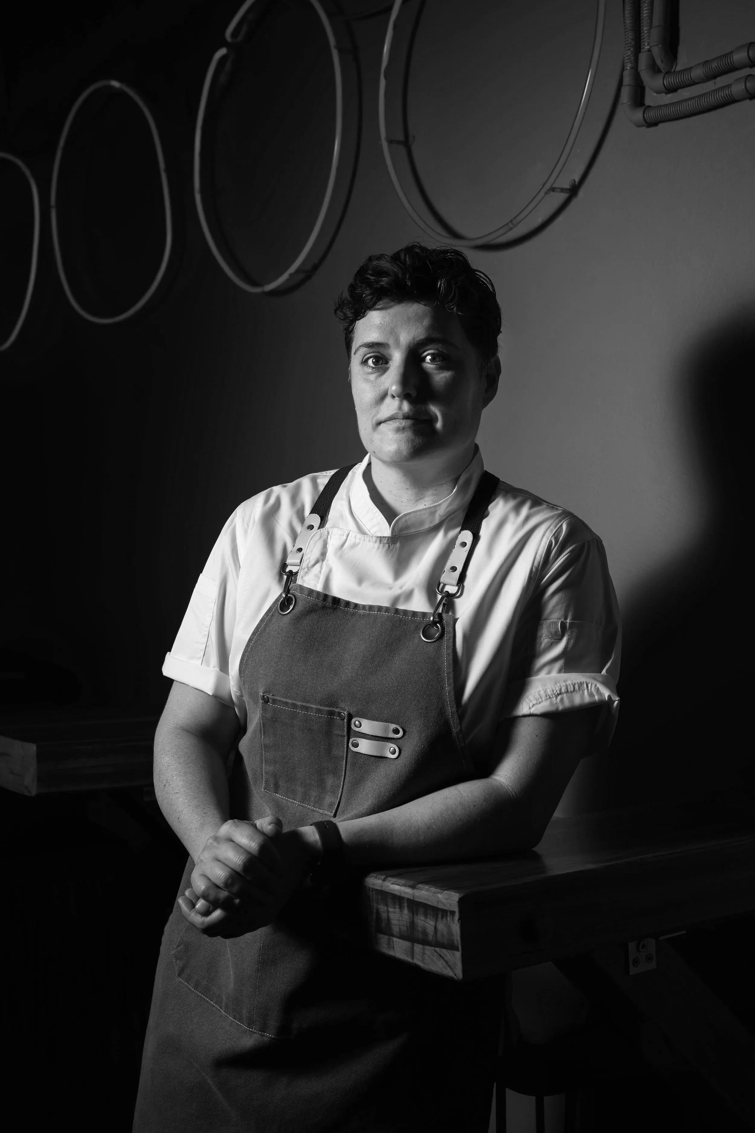 A black and white photo of a woman wearing a chef's coat and apron, standing behind a wooden counter with her hands clasped together, with a dark background and circular metal frames on the wall behind her.