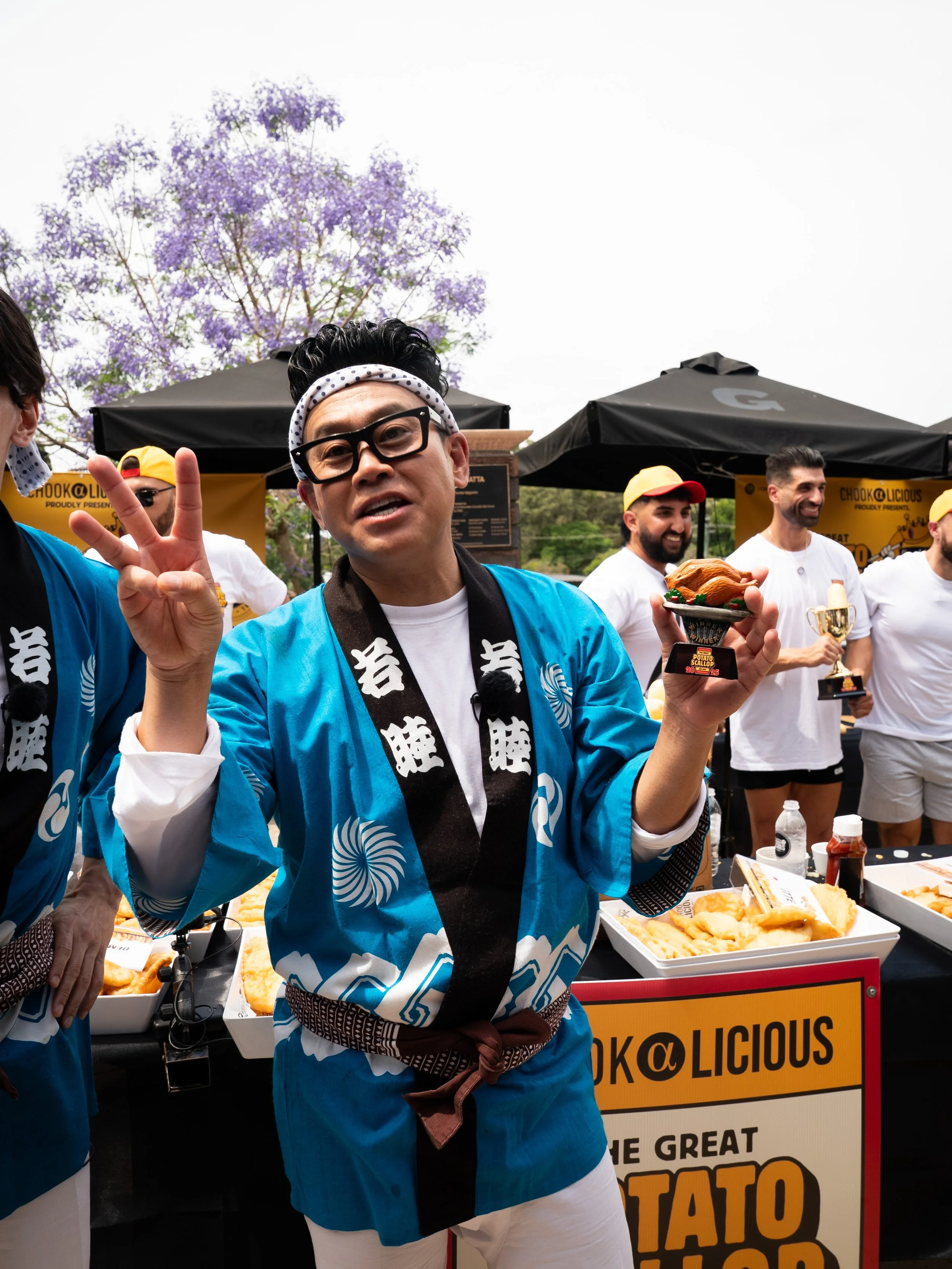 A man in a blue traditional Japanese festival shirt and glasses holding a trophy, making a peace sign, surrounded by people at an outdoor event with food stands and a purple flowering tree in the background.