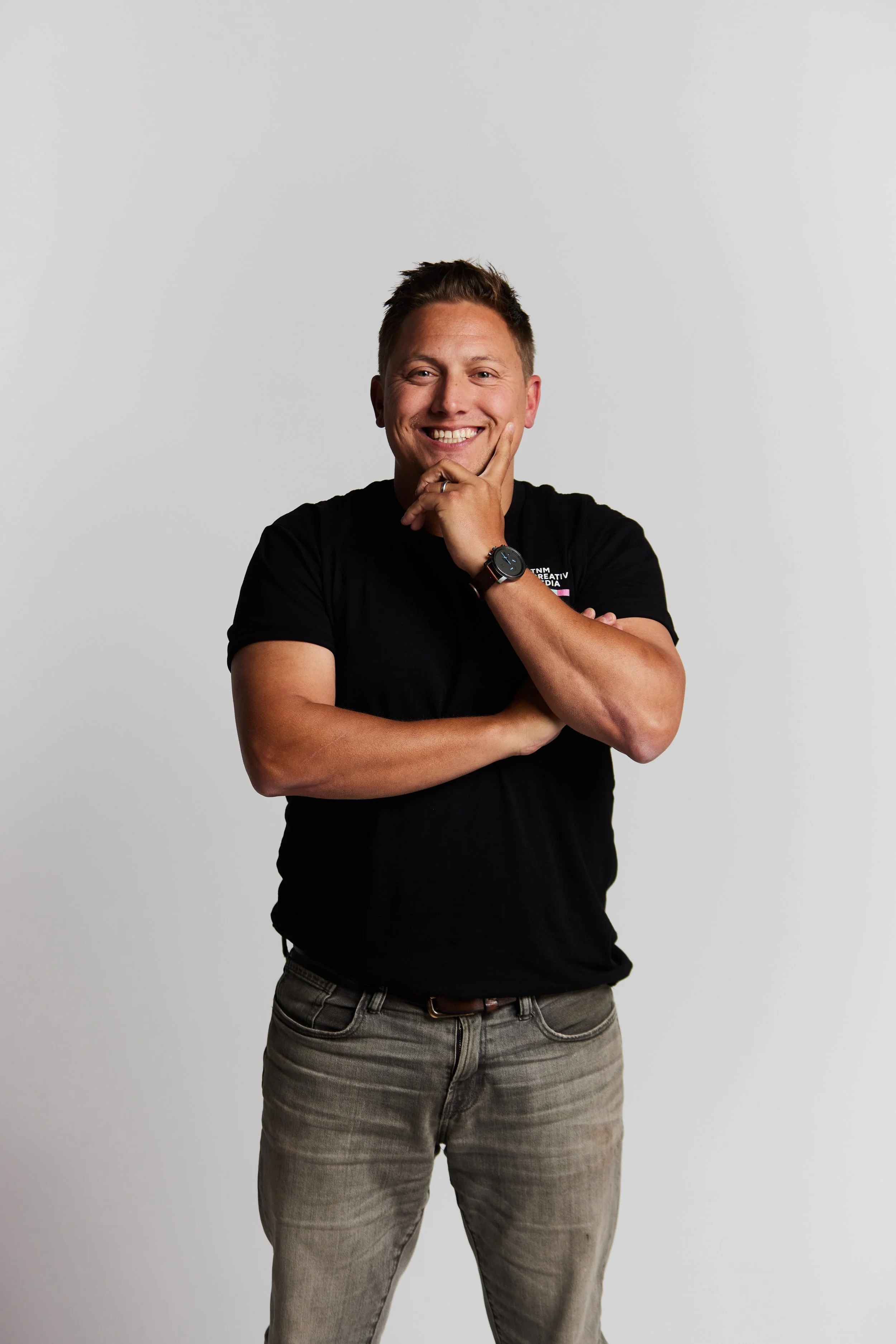 A man with short brown hair, wearing a black t-shirt and light gray jeans, smiling and posing with one arm crossed and the other hand touching his chin, against a plain white background.