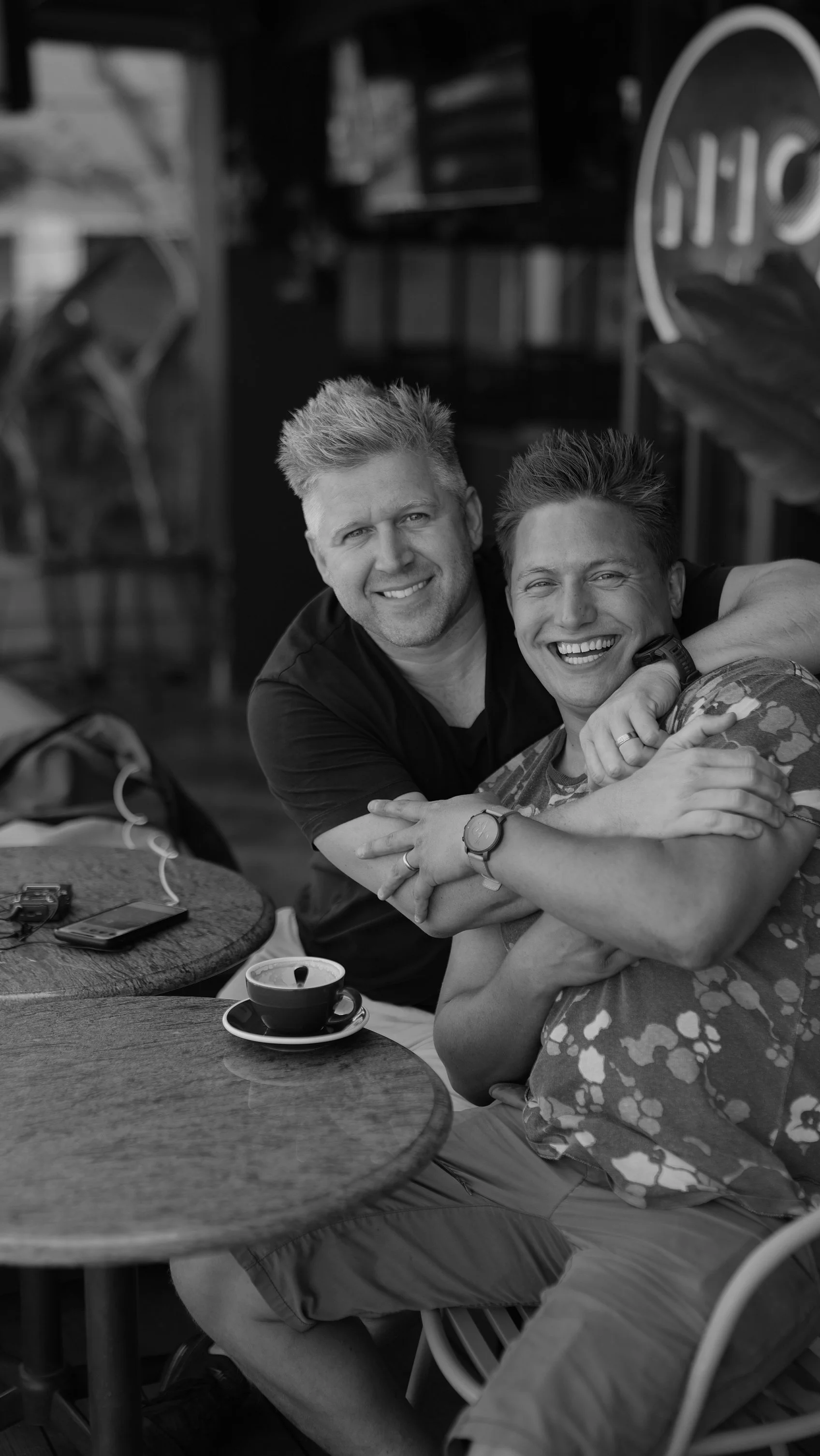 Two smiling men hugging at a table with a cup of coffee and a smartphone, in a cozy indoor setting.