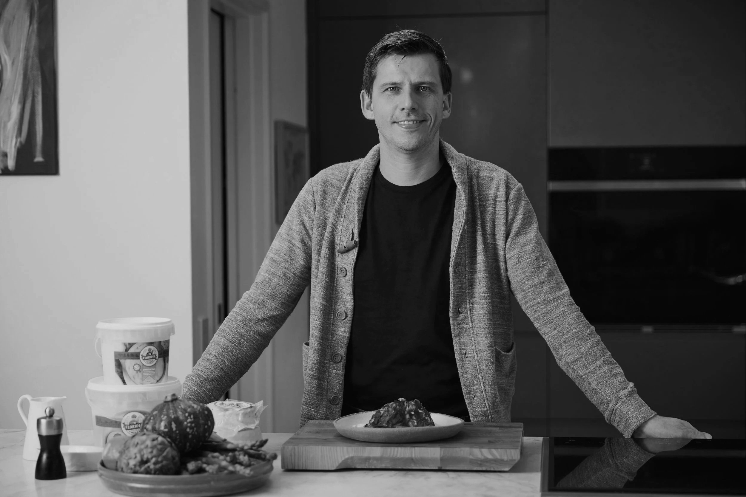 A man standing at a kitchen counter smiling, with a plate of food in front of him. There are jars and a pumpkin on the counter, with a background of modern kitchen cabinets.