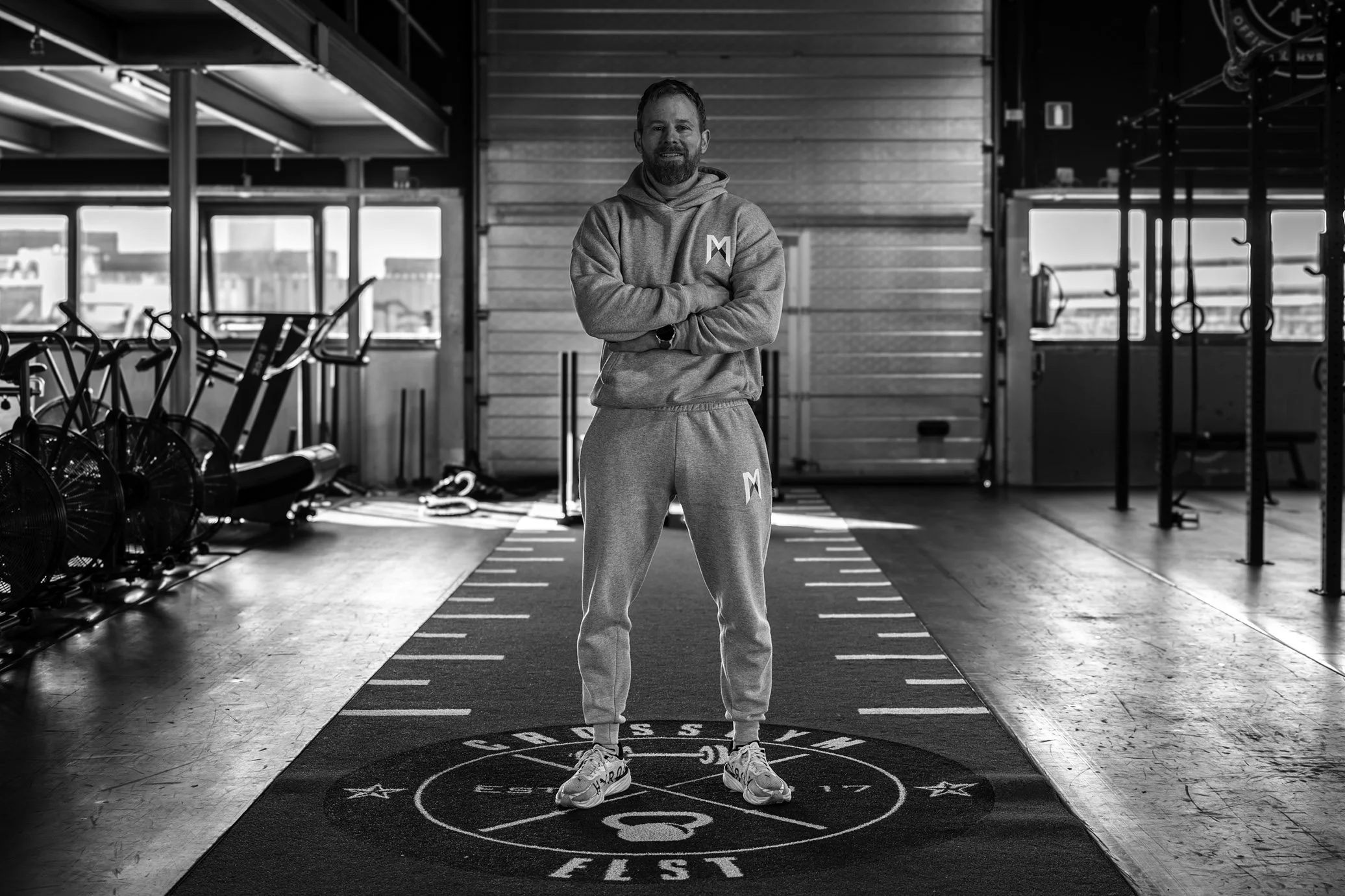 Een man in sportkleding staat met armen over elkaar in een sportschool, zwart-wit afbeelding, sportuitrusting zichtbaar op de achtergrond.