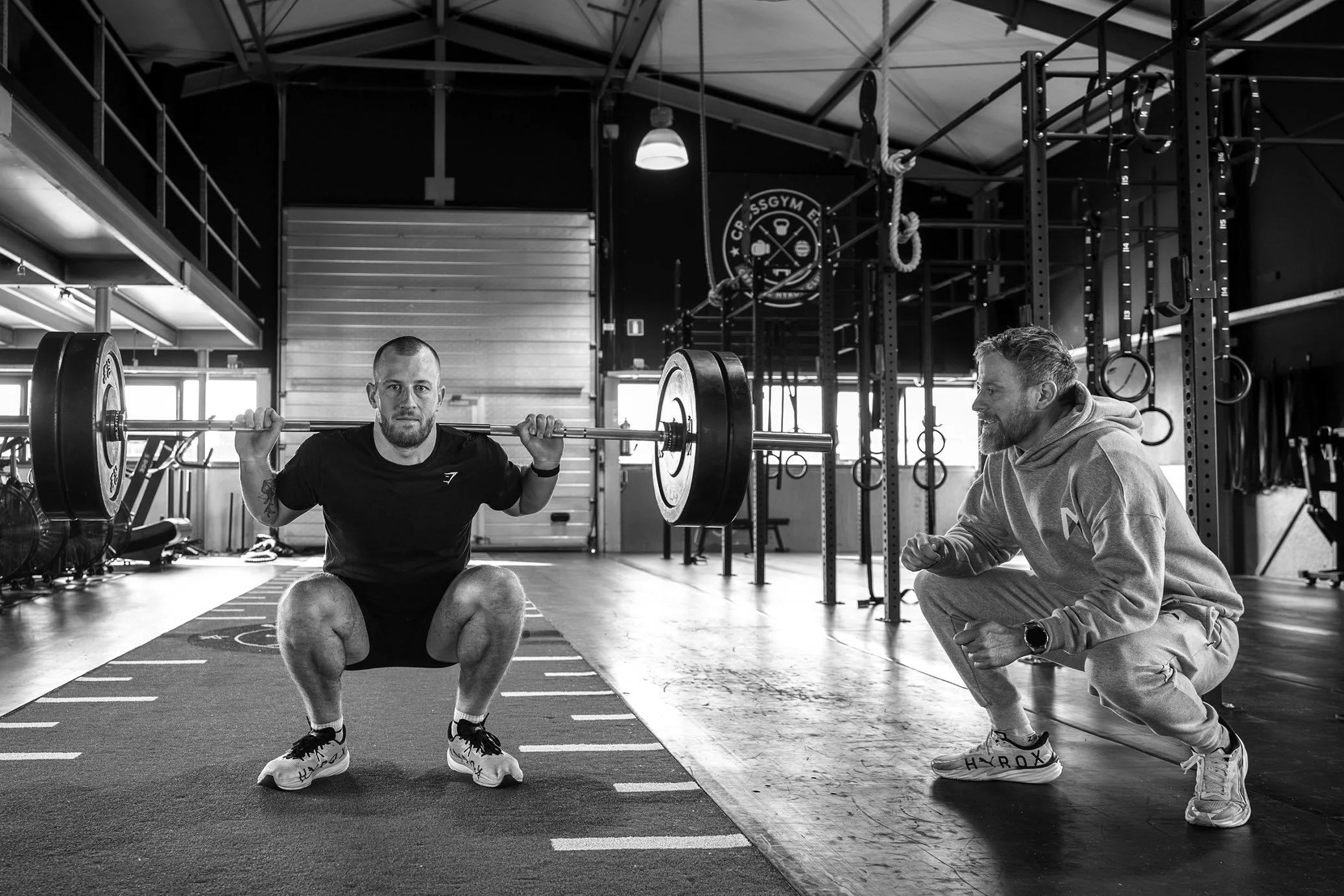 Man doet squats met een barbell onder toezicht van een trainer in een crossfit gym.