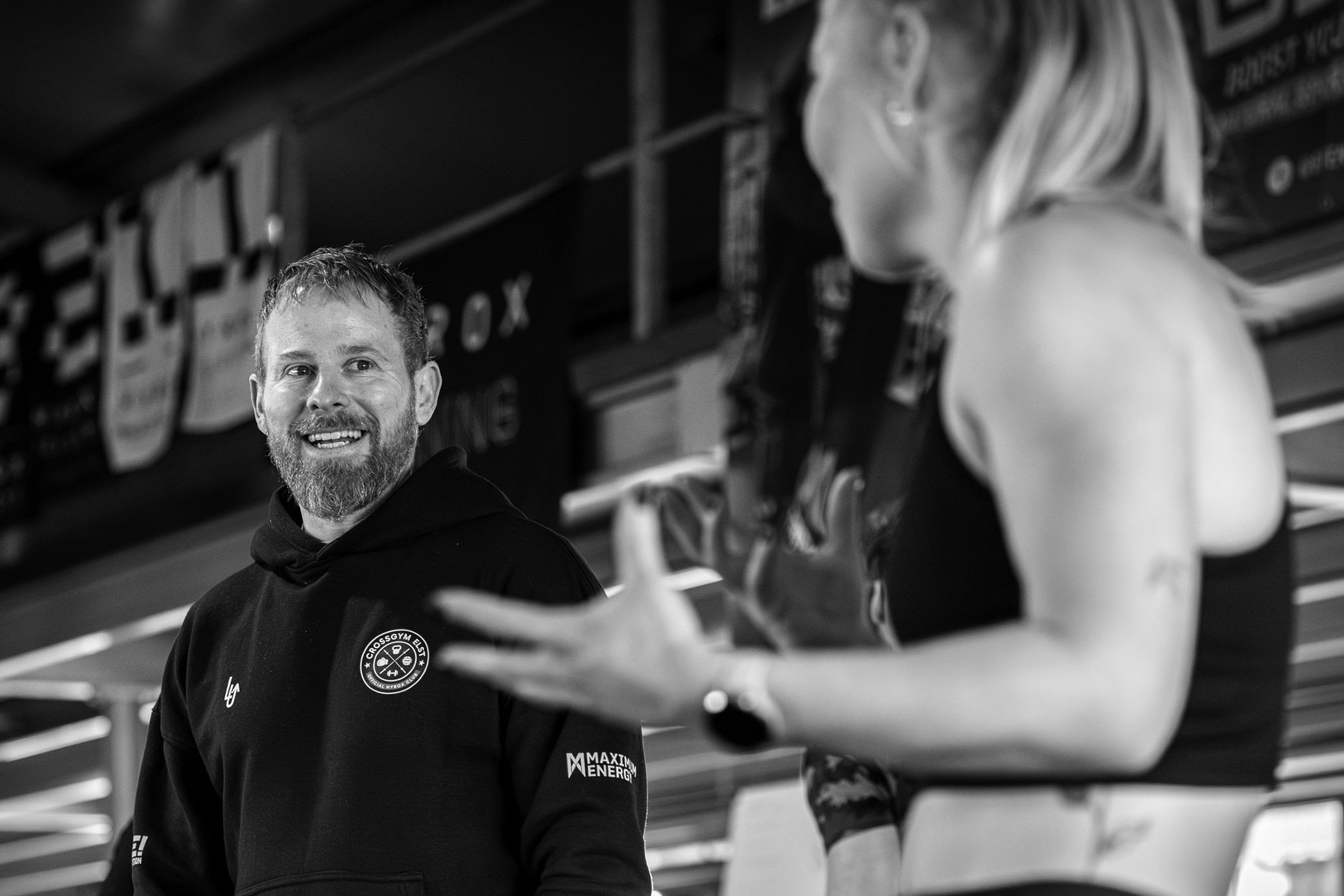 Een man met een baard en een glimlach draagt een zwarte hoodie met het logo van Crossgym EIJ en kijkt naar een vrouw die een sporttop en een fitness tracker aan heeft. Ze lijkt te praten in een sportschoolomgeving.