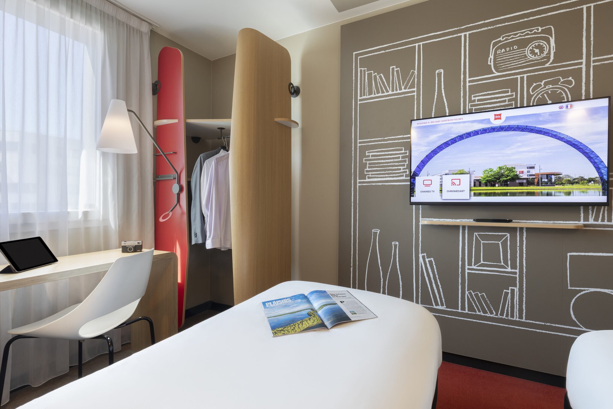 Interior de una habitación de hotel con un escritorio, una silla blanca, un televisor, una estantería empotrada con ropa, una pared decorada con dibujos discretos y una gran ventana con cortinas blancas. Sobre la mesa hay una revista abierta.