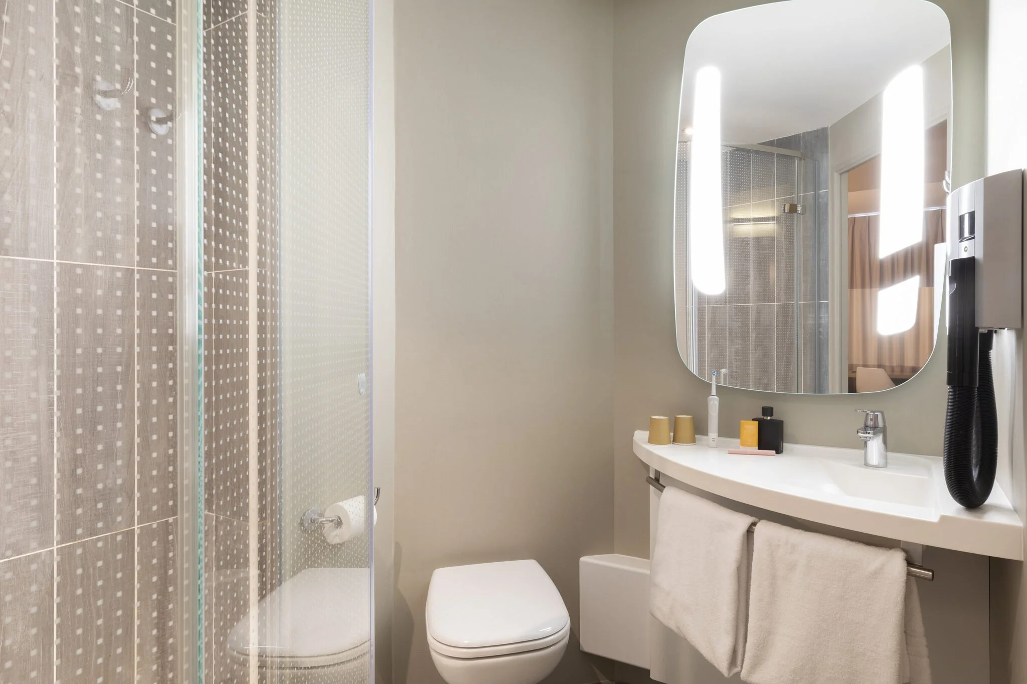 Un cuarto de baño moderno con un espejo rectangular sobre un lavabo blanco, un secador de pelo negro fijado a la pared, una toalla blanca colgada, vasos de papel, un jabón, un dispensador de jabón y un frasco de gel de baño sobre la encimera. El cuarto también cuenta con una cabina de ducha con puerta de cristal. La luz natural entra por una ventana de la estancia.