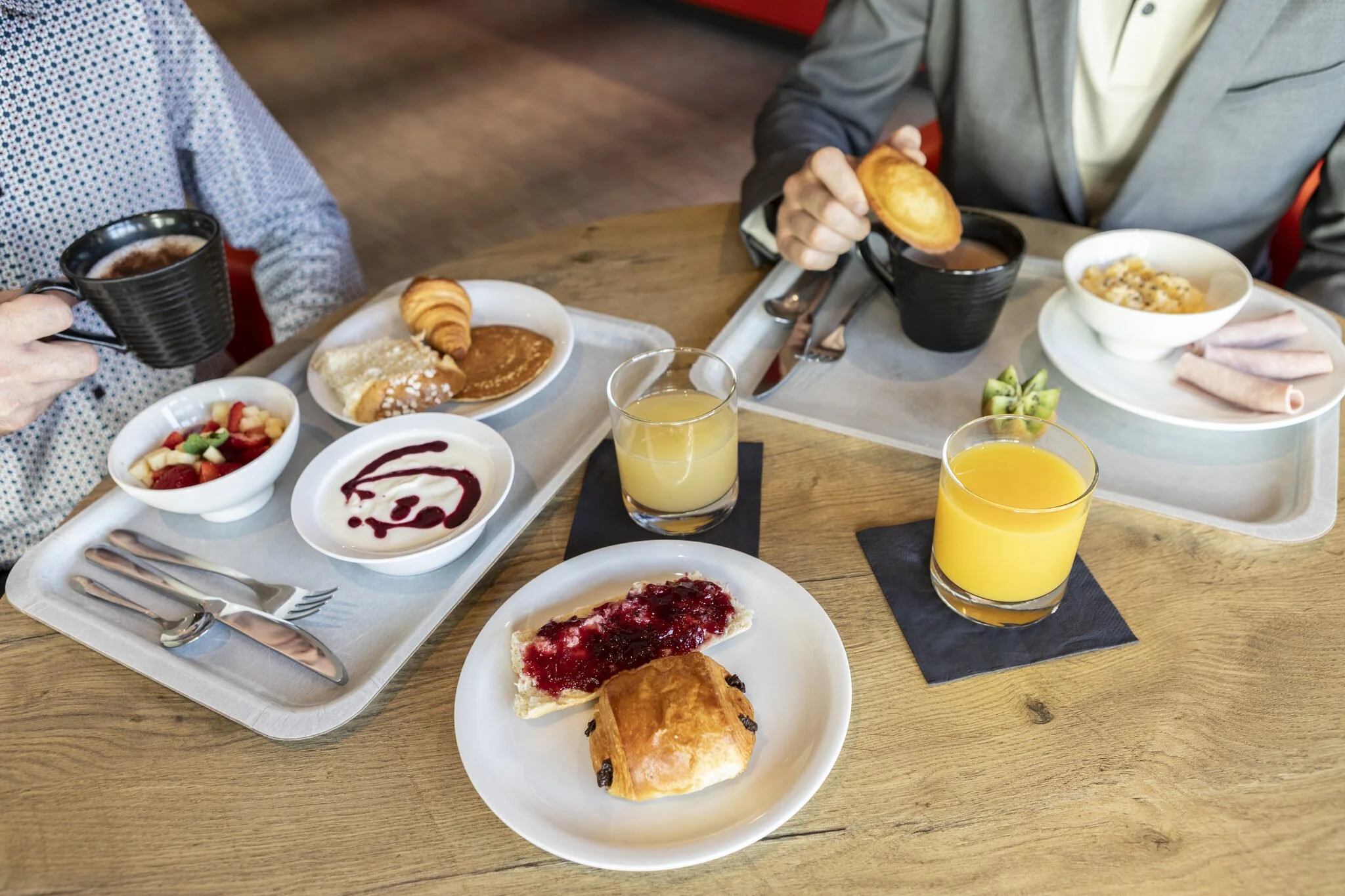 Breakfast consisting of pancakes, croissants, fruit, yogurt, sausages, and two glasses of orange juice, with two people drinking hot chocolate from mugs.