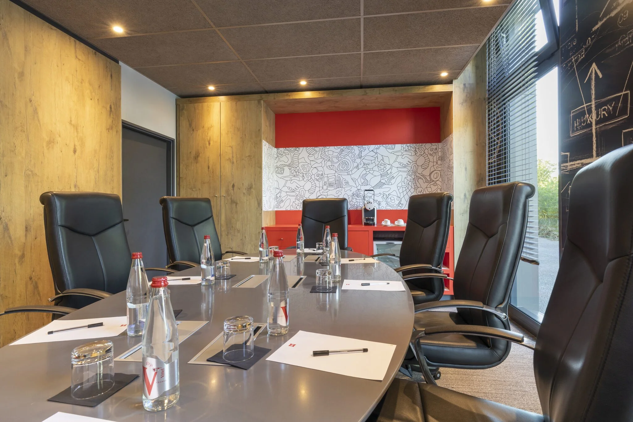 Sala de reuniones con una mesa, sillas de cuero negro, botellas de agua, blocs de notas y bolígrafos, paredes de madera, una pared con pizarra y ventanas con persianas, ambiente moderno