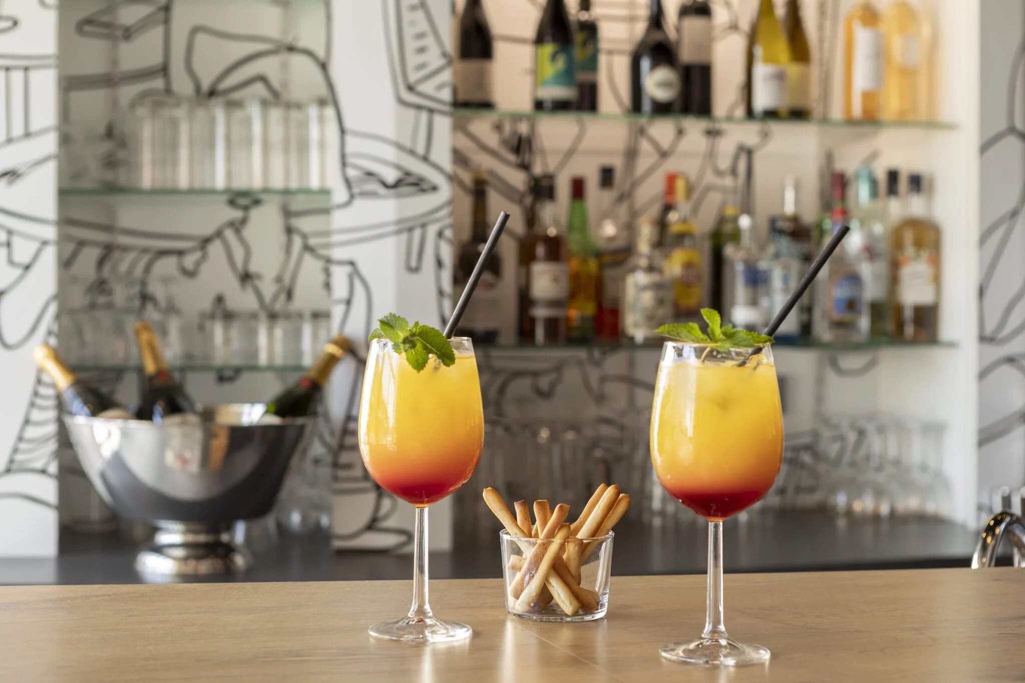 Dos cócteles de colores en vasos de cóctel con hojas de menta, acompañados de palitos en una botella de cristal, con una barra de fondo.
