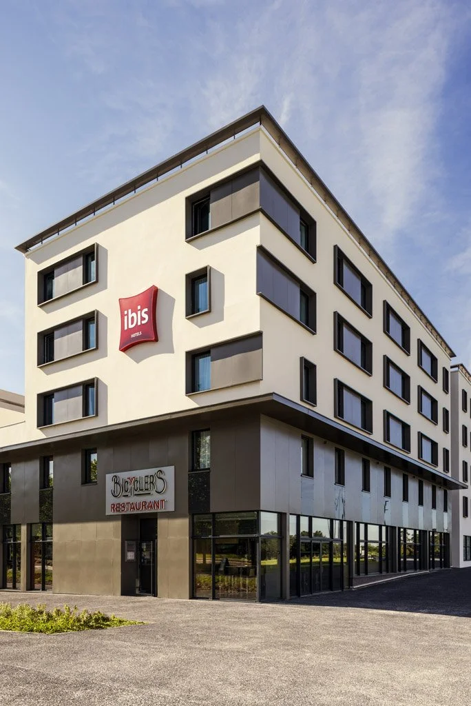 Modern multi-story Ibis hotel building with parked area in front, clear sky