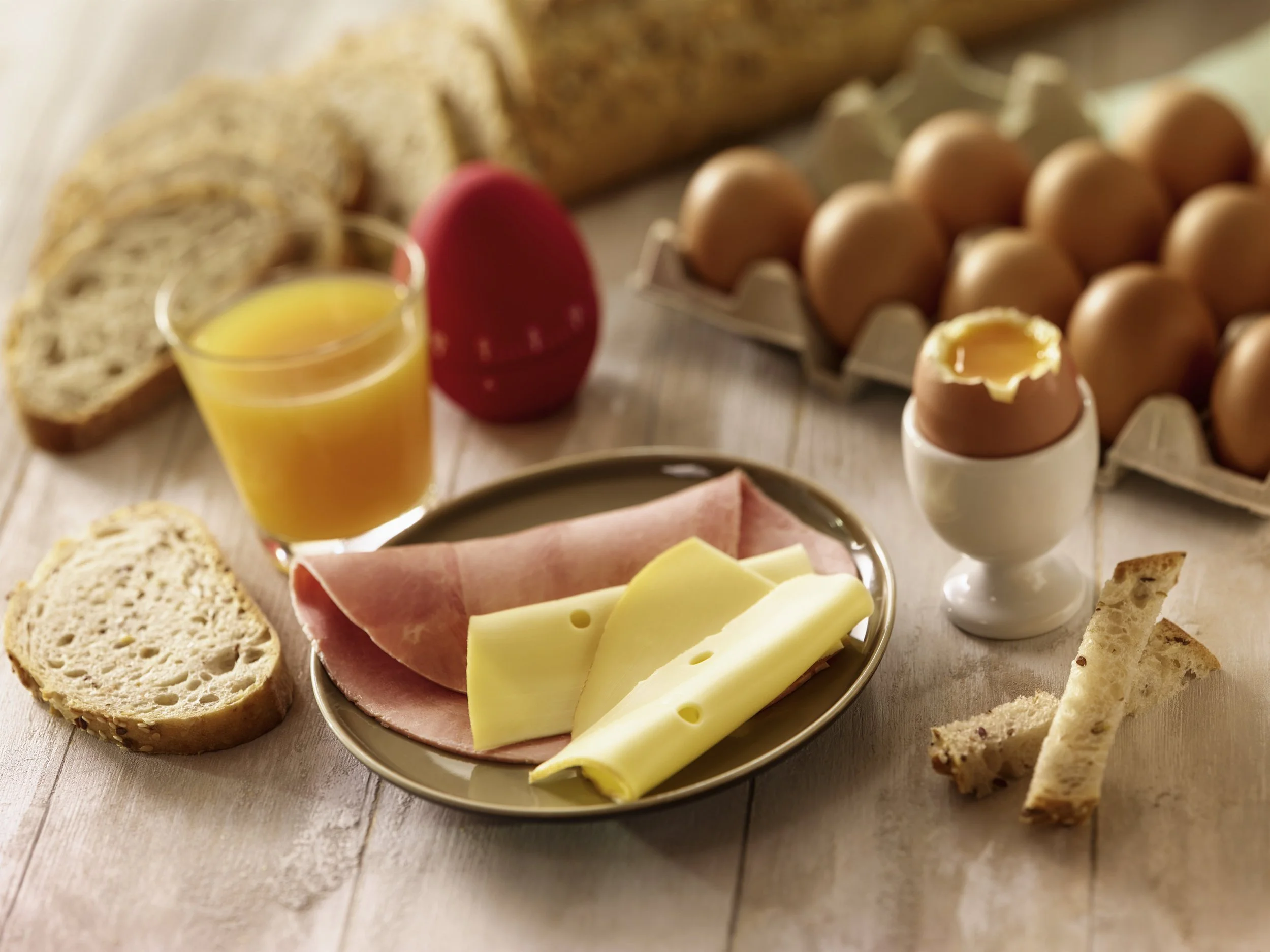 Breakfast assortment including sliced bread, a glass of orange juice, a red egg-shaped container, a tray of eggs, a soft-boiled egg in an egg cup, sliced ham, sliced cheese, and breadsticks on a wooden surface.