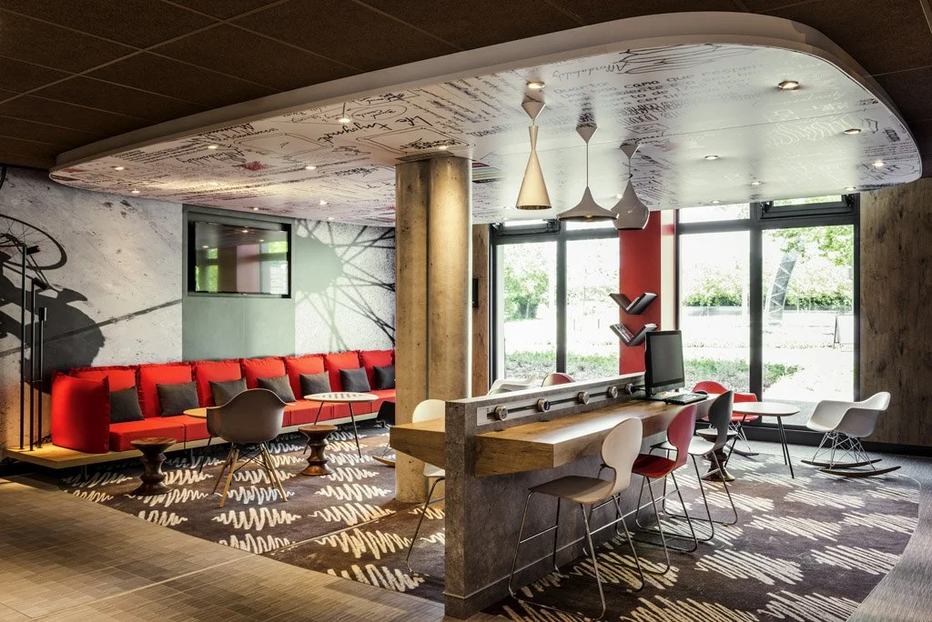Modern lounge area with a curved ceiling, large windows, red and gray seating, a wooden reception counter with chairs, and decorative pendant lights.