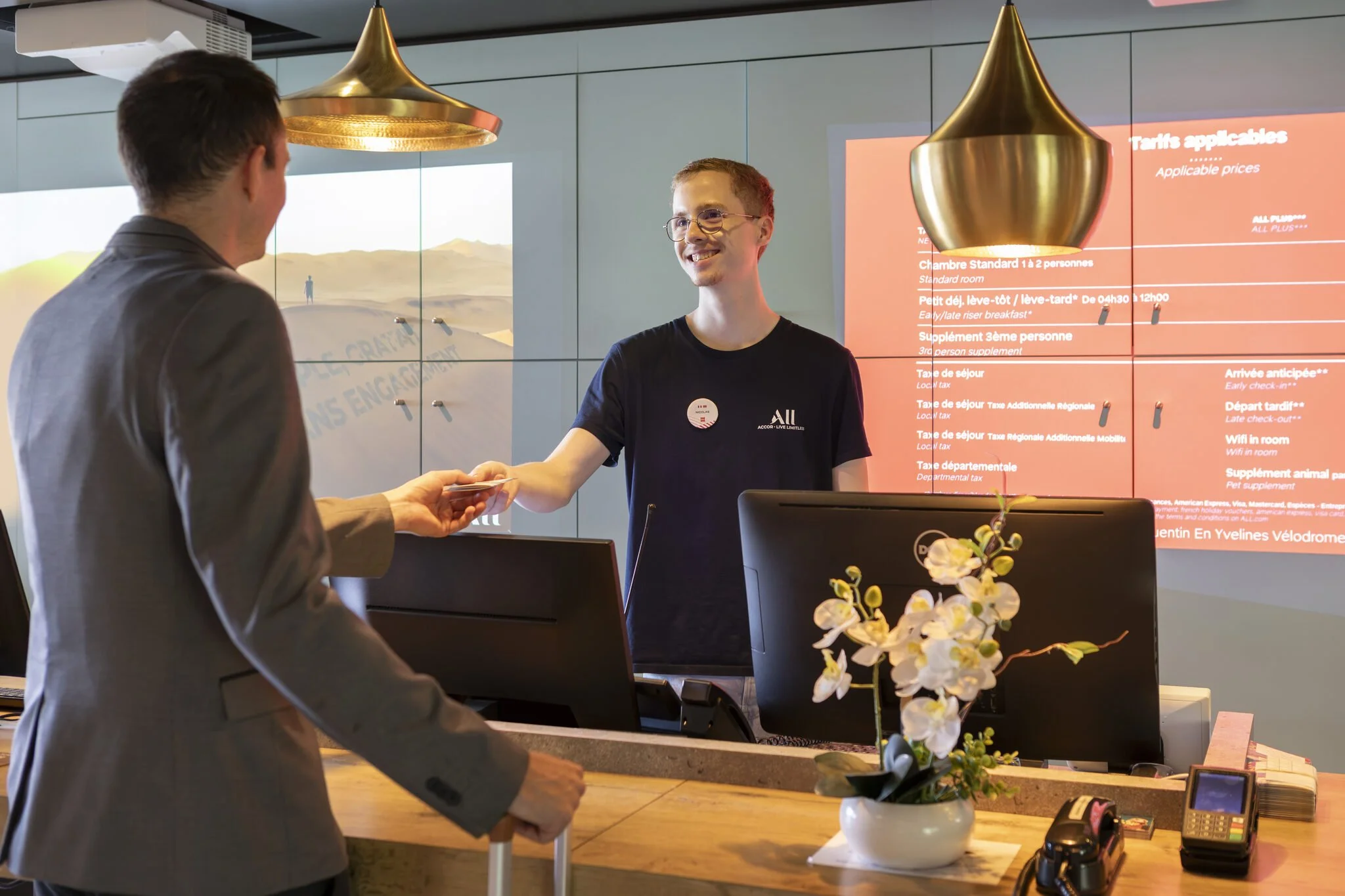 Un employé de comptoir en train de remettre une carte à un client à la réception d'un hôtel, avec un panneau d'informations sur l'écran derrière eux.