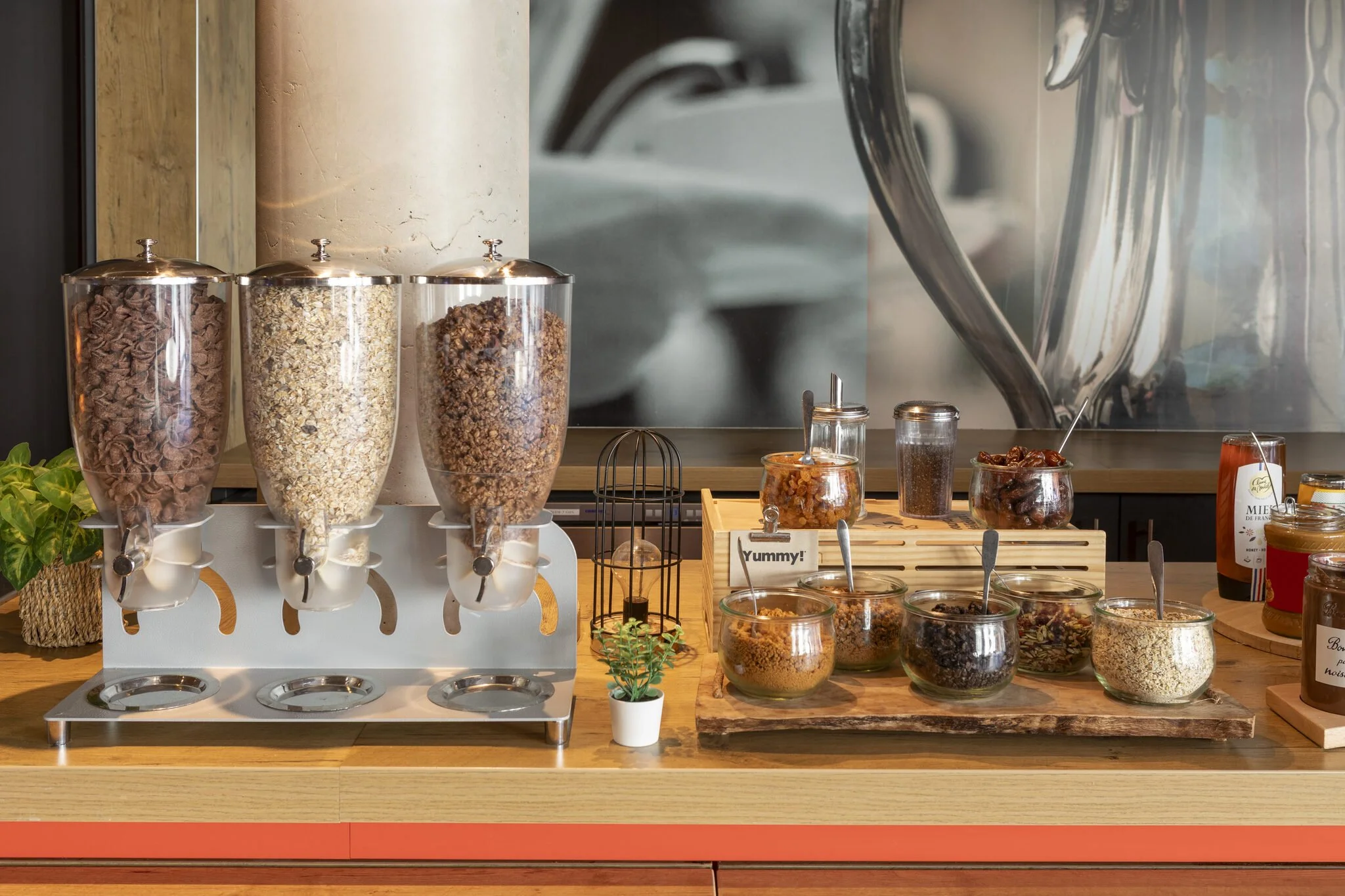 A display case featuring various types of cereal in glass containers, along with jars of jam, a small frame with the word "Yummy!" and a houseplant, set in a café or grocery store setting.
