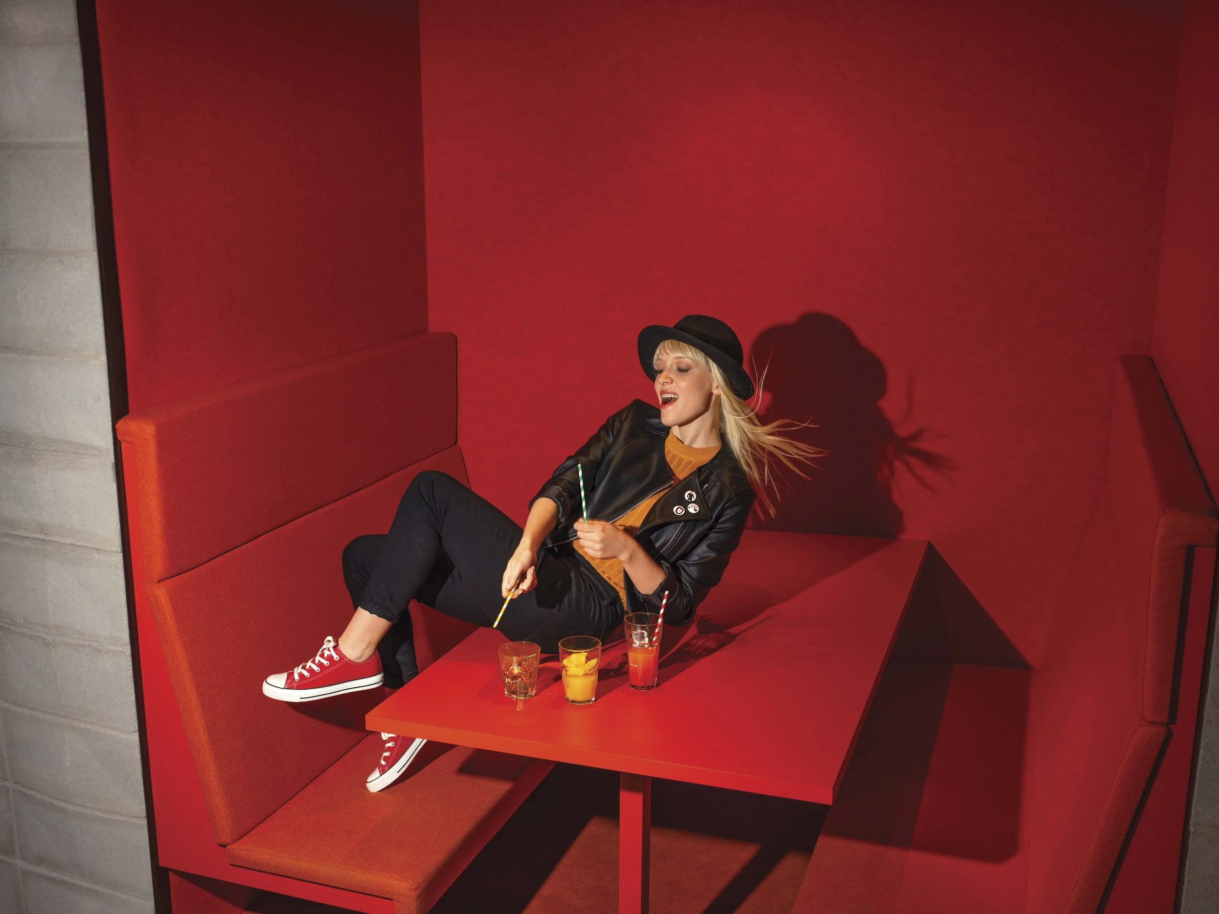 A young woman with blonde hair sitting in a red booth, wearing a black hat and leather jacket, smiling with her eyes closed, holding a green striped straw, with three colorful drinks on the red table in front of her.