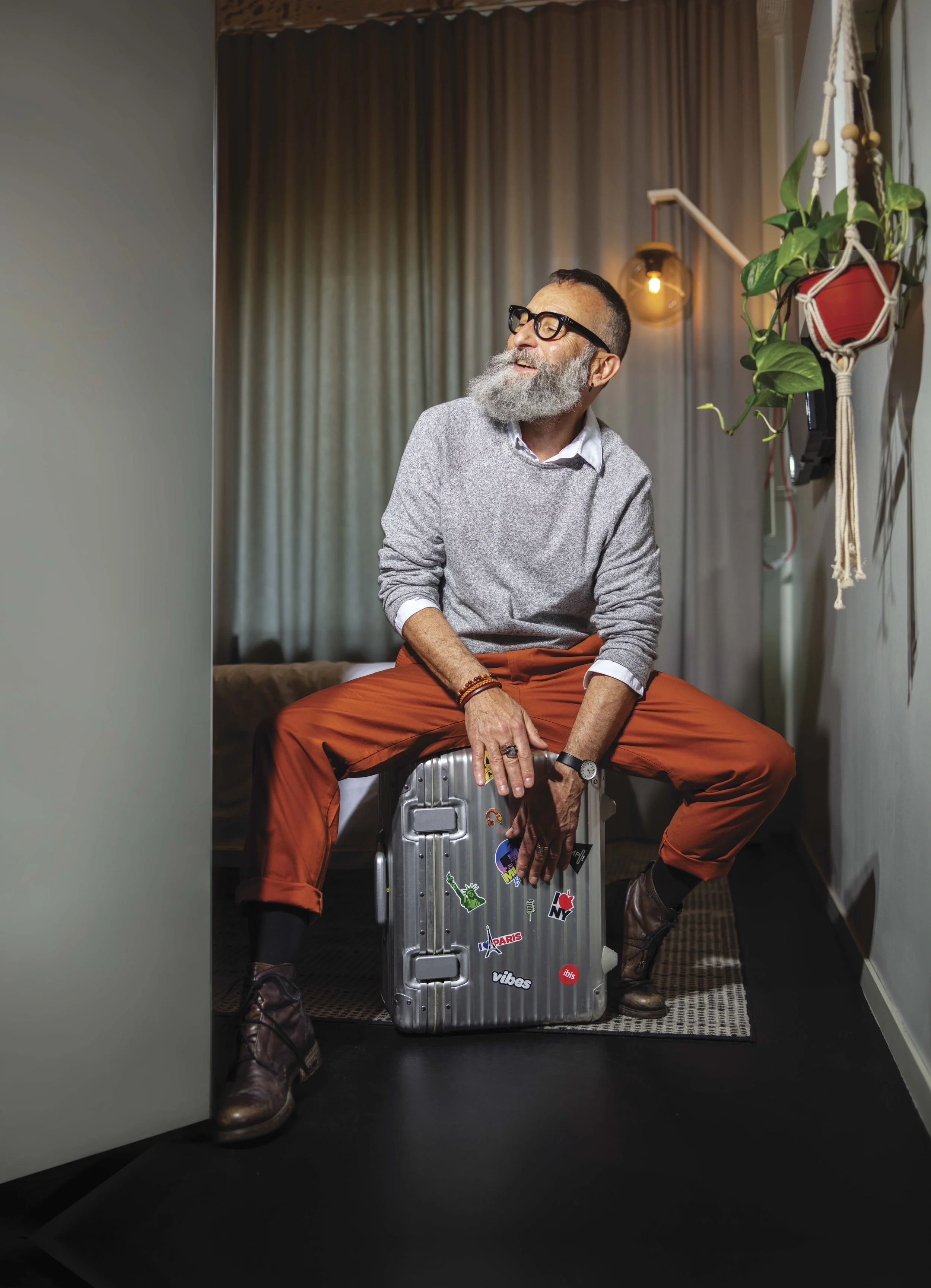 A man with a beard and glasses, wearing an argyle sweater, sitting on a suitcase decorated with travel stickers, in a doorway of a room with curtains and a hanging light fixture, smiling and looking away.