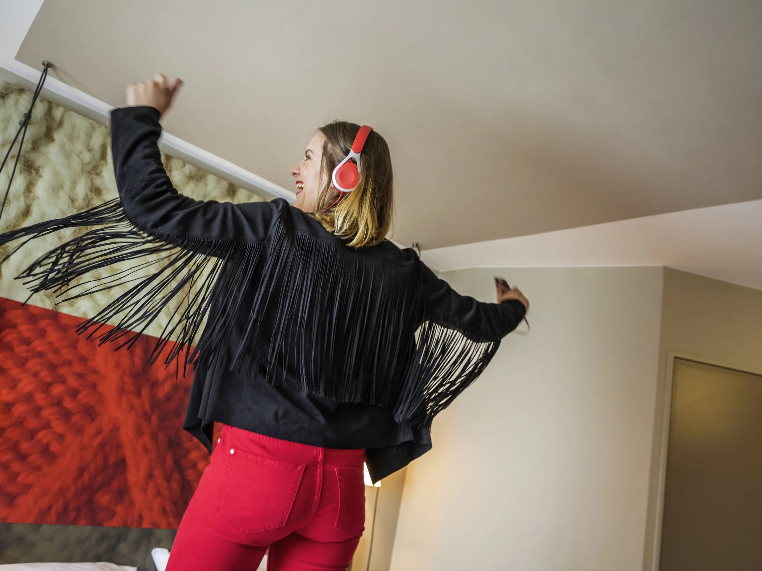 A woman with shoulder-length hair wearing red pants and a black jacket with fringes, standing indoors next to a red textured wall and a beige wall, smiling and holding onto a railing with her left hand at Ibis Saint quentin en yvelines.