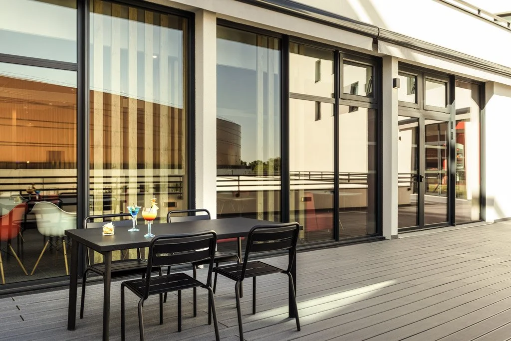 Outdoor patio with a black table and three black chairs in front of large glass windows and doors of a building, with drinks on the table.