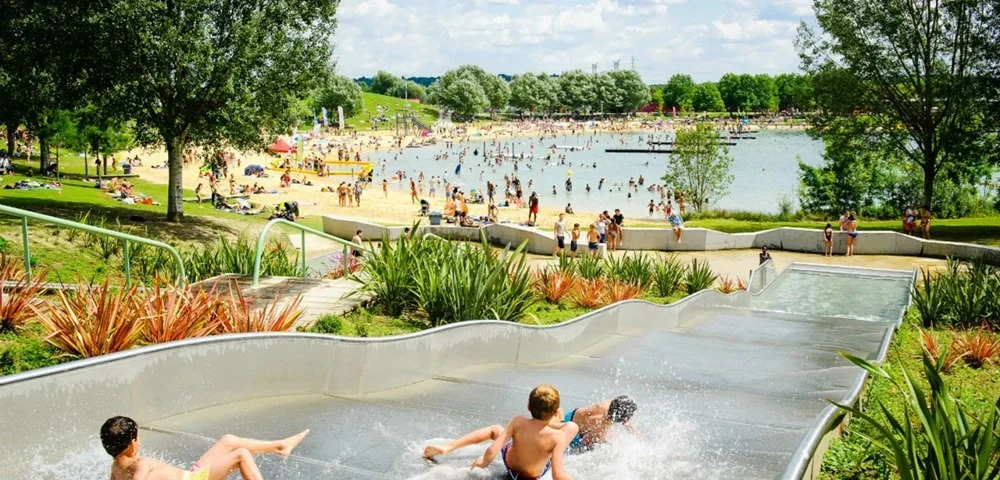 People enjoying a public water park with a lazy river, surrounded by trees and greenery on a sunny day.