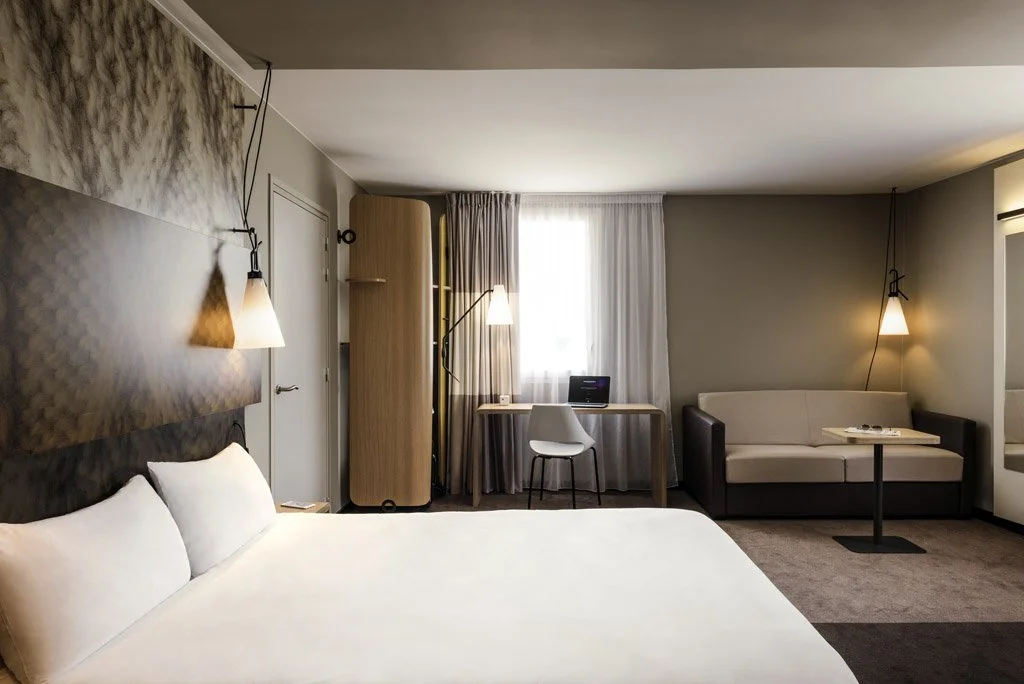 Hotel room with a bed, sofa, desk, and reading lamps, featuring simple decor and neutral colors.