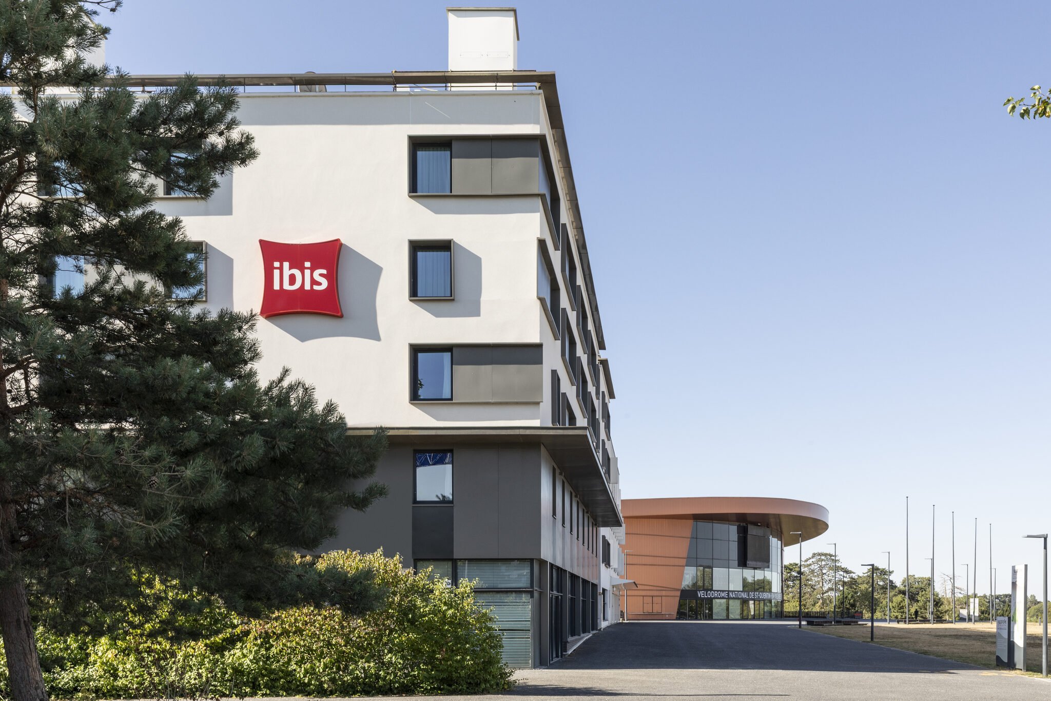 Une grande bâtisse moderne au design épuré avec un logo 'ibis' rouge sur la façade. Il y a un arbre à gauche de l'image et un ciel clair en arrière-plan.