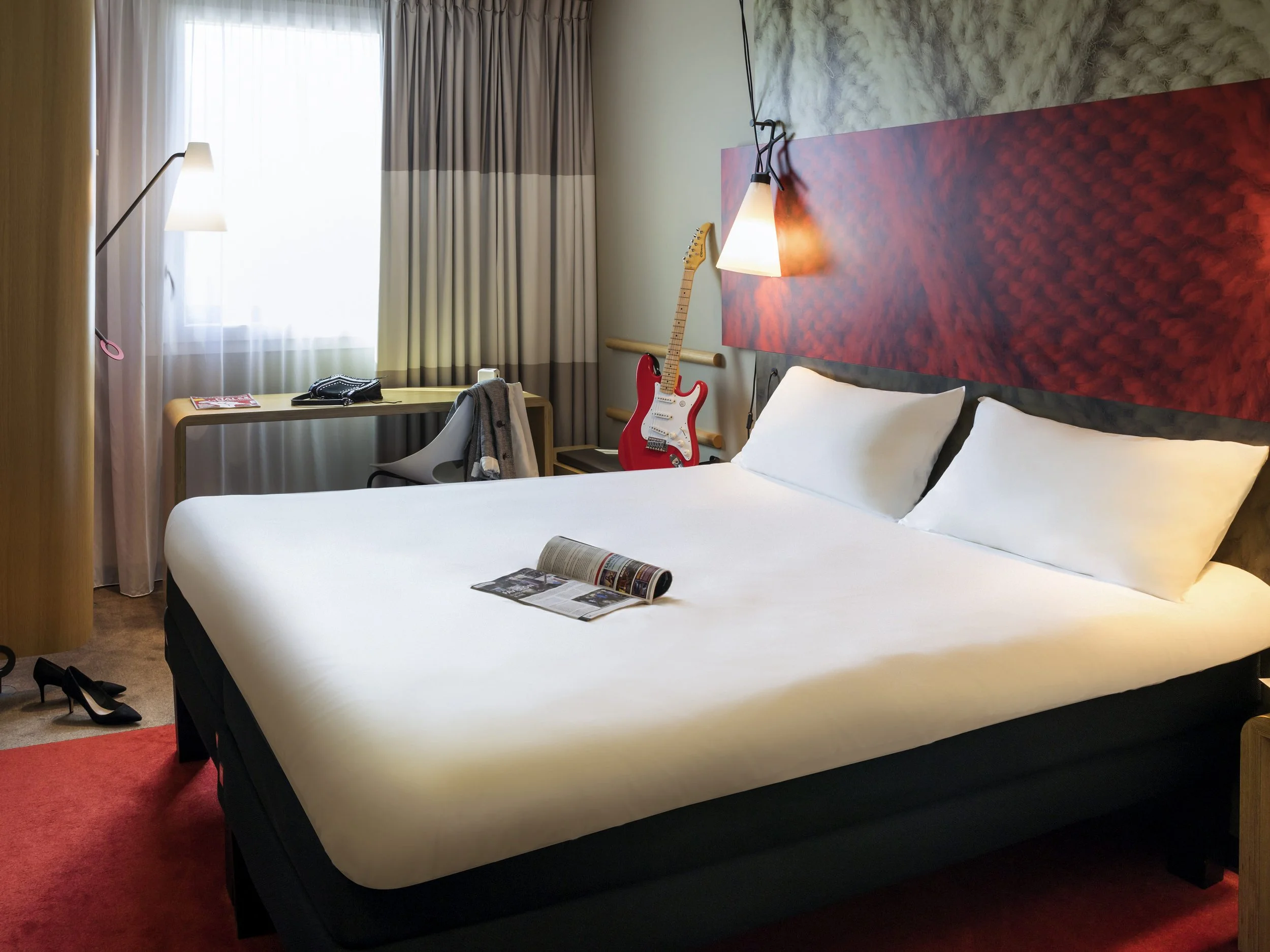 Hotel room with a bed, a guitar, a desk with a chair, and a pair of black high-heeled shoes on the floor.