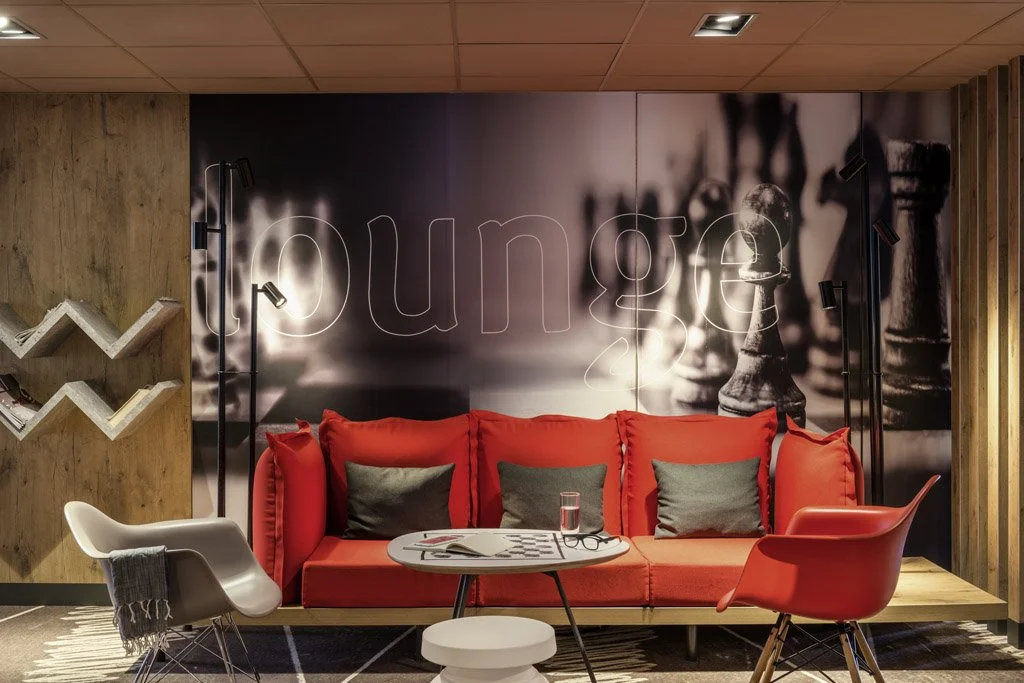 Modern lounge area with a red sofa, black and gray cushions, a round coffee table, a white armchair with a gray throw blanket, a red chair, and a wall mural with the word 'lounge' and chess pieces in black and white.