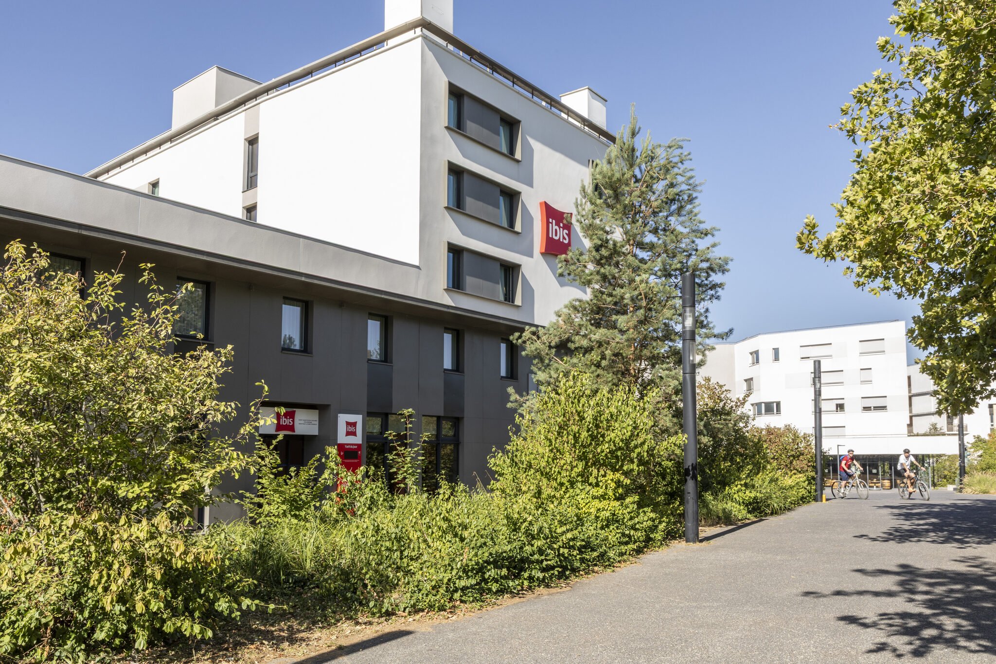 Un edificio del hotel Ibis con ventanas modernas, rodeado de árboles verdes y bajo un cielo azul claro en un día soleado. Varias personas van en bicicleta o caminan por una acera junto a la fachada.