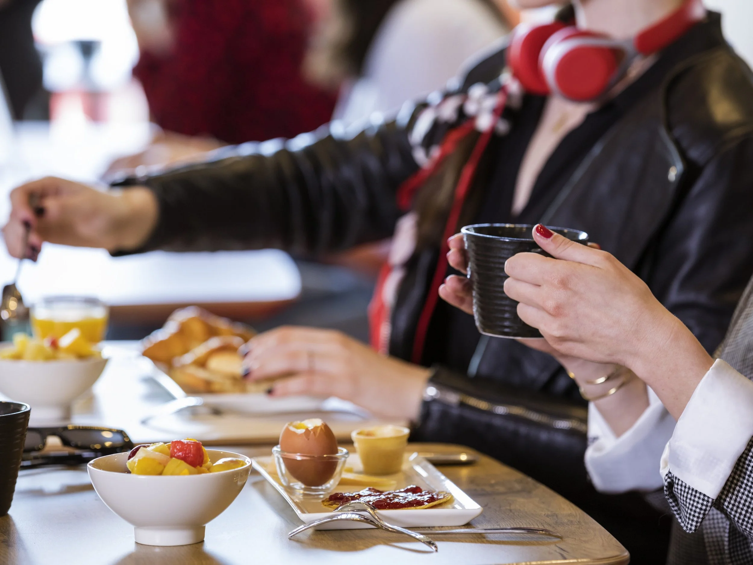 People enjoying breakfast at a table with bowls of fruit, eggs, and toast, with one person holding a cup and another reaching for food.
