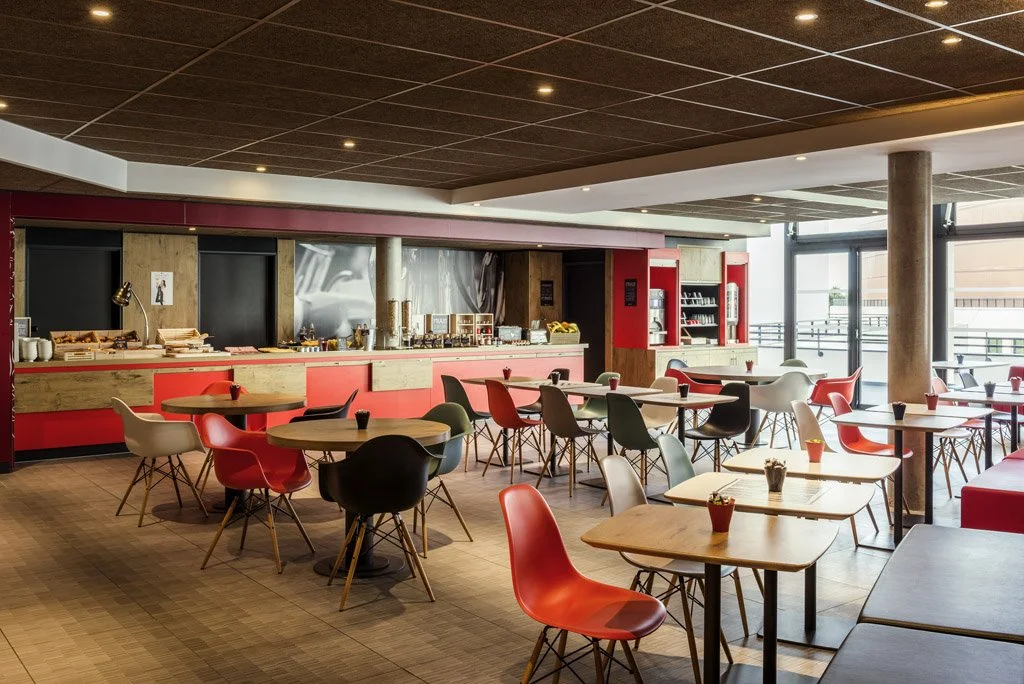 Modern breakfast area with round and square tables, colorful chairs, a food buffet, and large windows letting in natural light.
