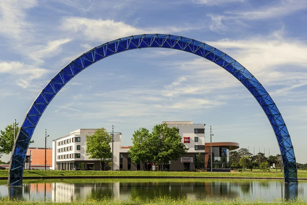 A large blue arch structure over a pond with a building and trees in background.
