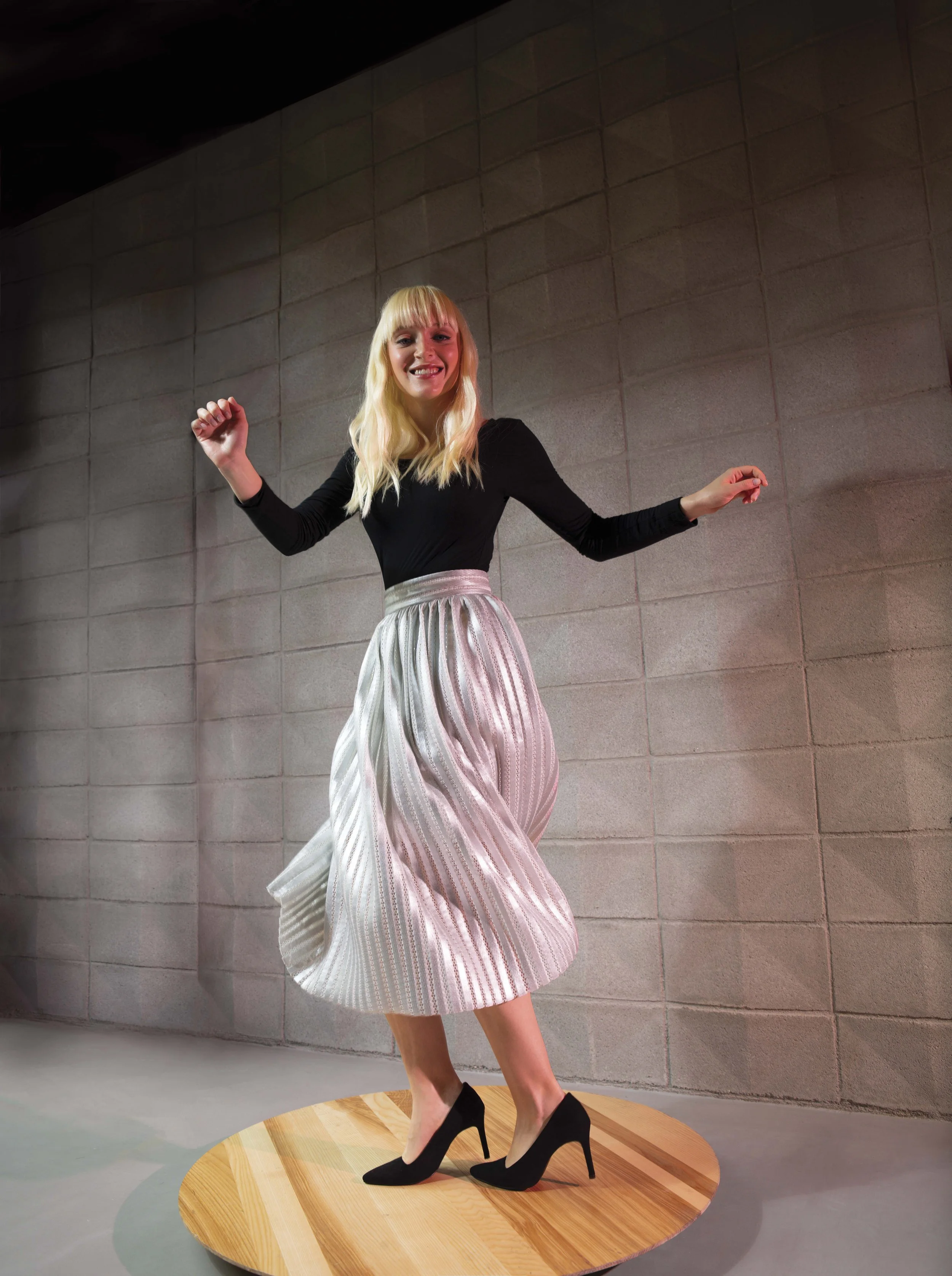 A woman with blonde hair and a black top dancing on a wooden platform with a striped skirt, against a gray cinderblock wall.