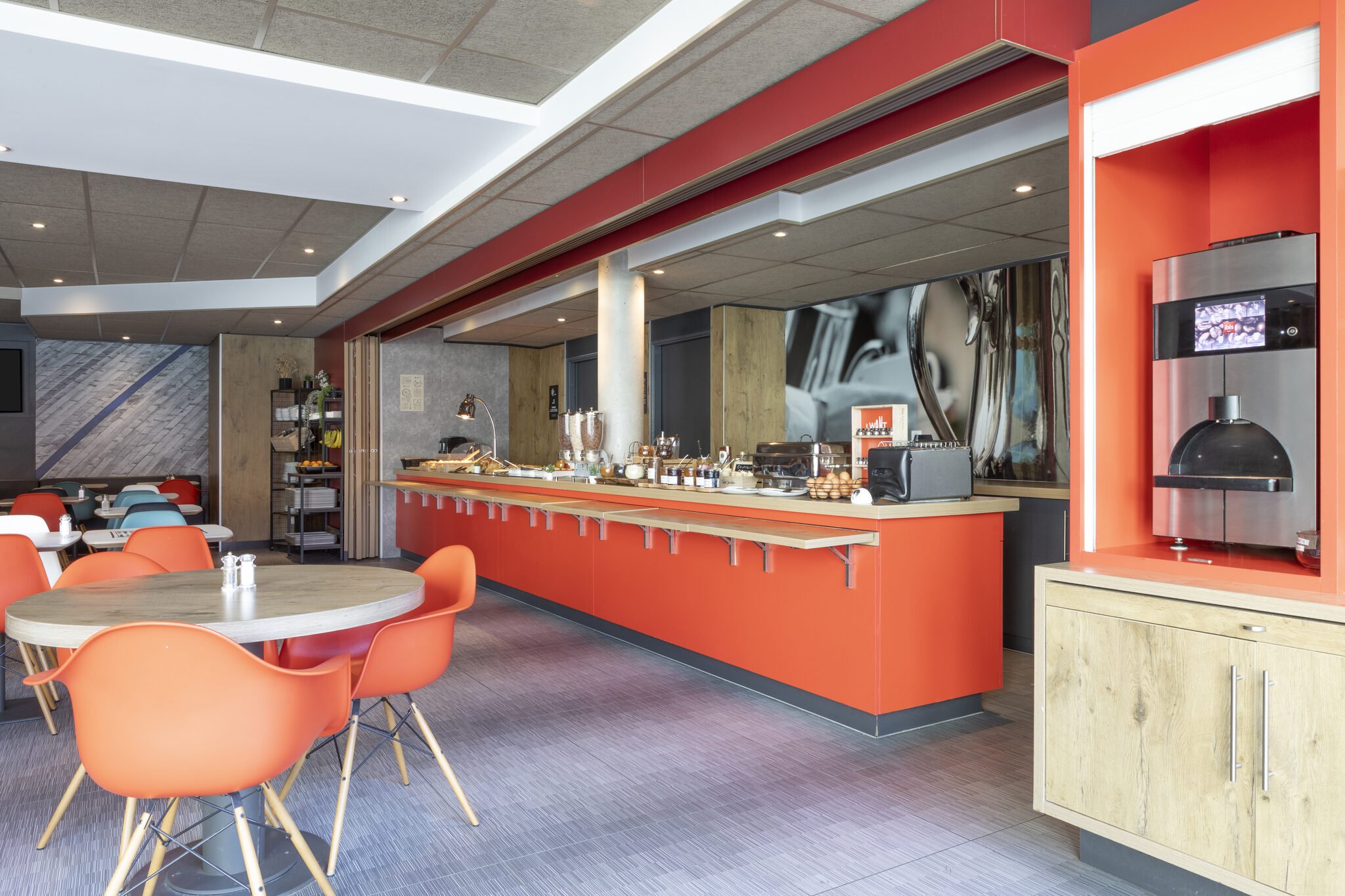 Interior de un restaurante con una barra de servicio roja, varias mesas redondas con sillas naranjas y una decoración moderna con una pared de madera y otra de piedra.