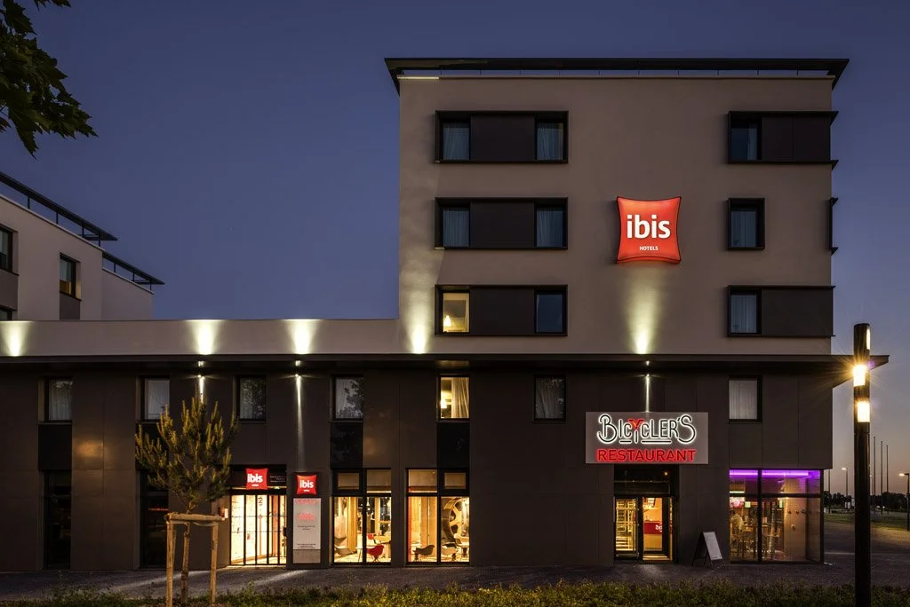 Exterior Ibis hotel building with lit signs for Ibis Hotel and a restaurant called Bicycler's, during dusk or evening.
