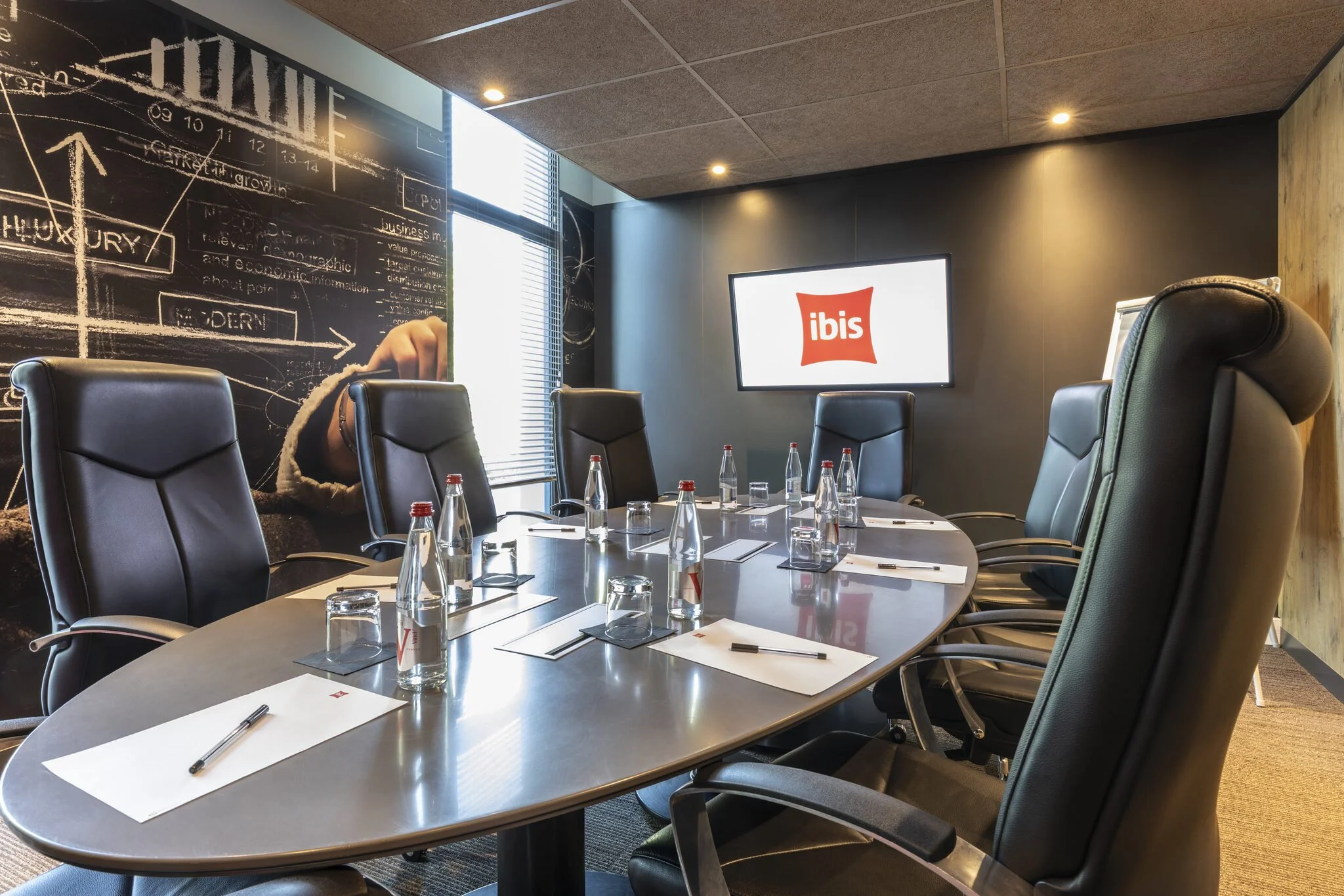 Salle de réunion moderne avec une grande table entourée de fauteuils, des bouteilles d'eau et des bloc-notes. Sur le mur, un écran avec le logo Ibis, et une peinture murale en noir avec des croquis et des diagrammes.