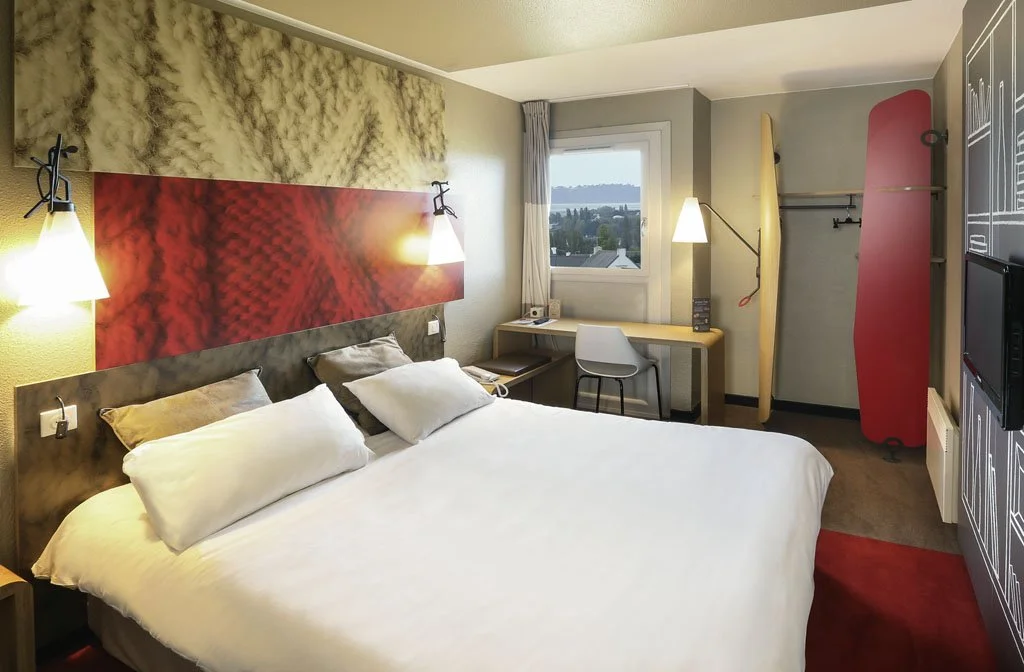 Hotel room with a bed, wall art, bedside lamps, a desk with a chair, a window showing a view, a skateboard, a wakeboard, and a red rug.