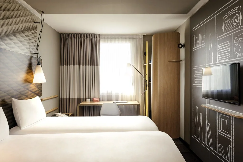 A modern hotel room with two beds, a wall-mounted TV, a desk with a chair, a wardrobe, and decorative chalkboard-style wall art.