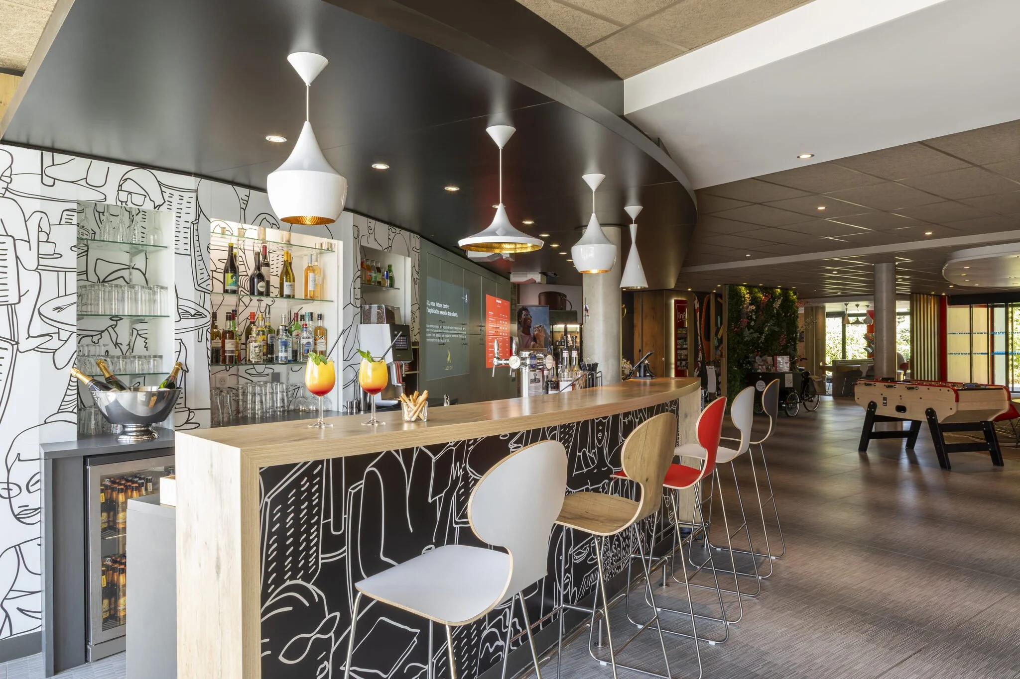 Un bar moderno con una barra de madera clara, varias sillas blancas y rojas, varias lámparas colgantes blancas y una pared decorada con dibujos minimalistas. También se aprecia una zona con juegos de mesa y sillones.